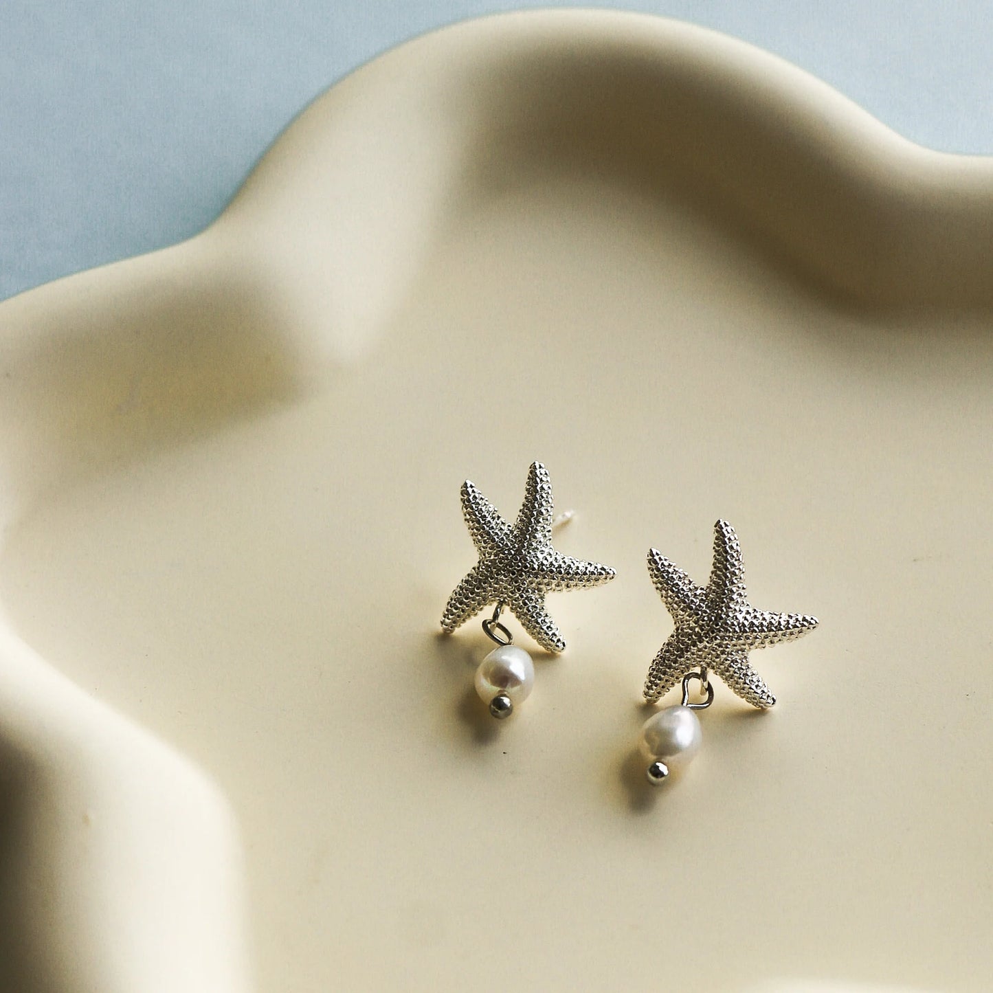 Boucles d'oreilles en forme d'étoiles de mer texturées avec perles d'eau douce. La paire est faite en laiton plaqué argent clair. Les boucles d'oreilles sont déposées sur une assiette en céramique beige sur une table grise. Plan éloigné sur les boucles d'oreilles.