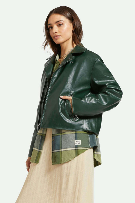 Manteau type perfecto en simili cuir lisse vert forêt porté par une femme. Le manteau est superposé sur une chemise et une robe longue. Vu de profil..