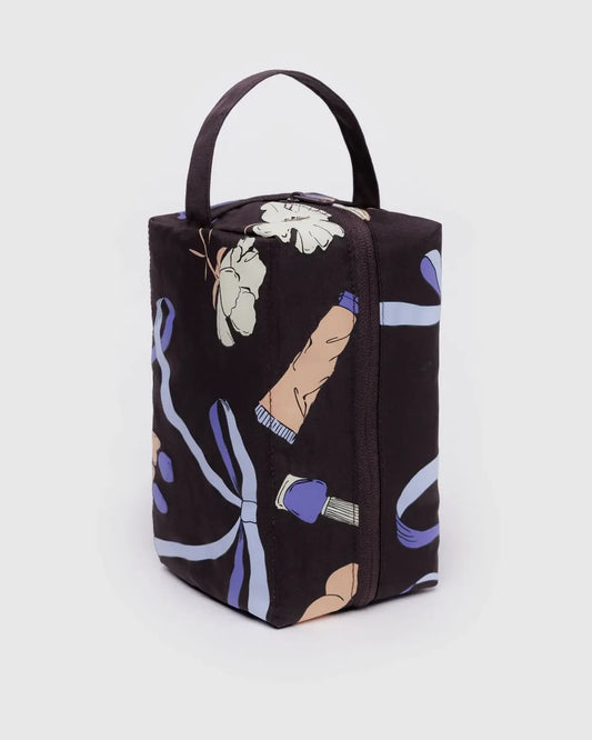 Trousse de toilette rectangulaire en tissu noir avec un motif illustré de rubans violets, fleurs et accessoires de beauté. Elle dispose d’une fermeture éclair sur le dessus et d’une anse intégrée pour un transport facile. Son design spacieux permet de ranger et organiser produits de beauté, maquillage ou articles de toilette. Idéale pour les voyages ou un usage quotidien.