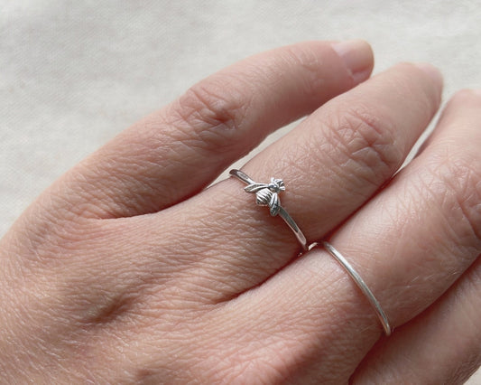 Bague délicate sur laquelle est soudée un petite abeille en argent sterling. L’anneau est une fine bande en argent martelé. La bague est porté à l’annulaire gauche d’une personne.