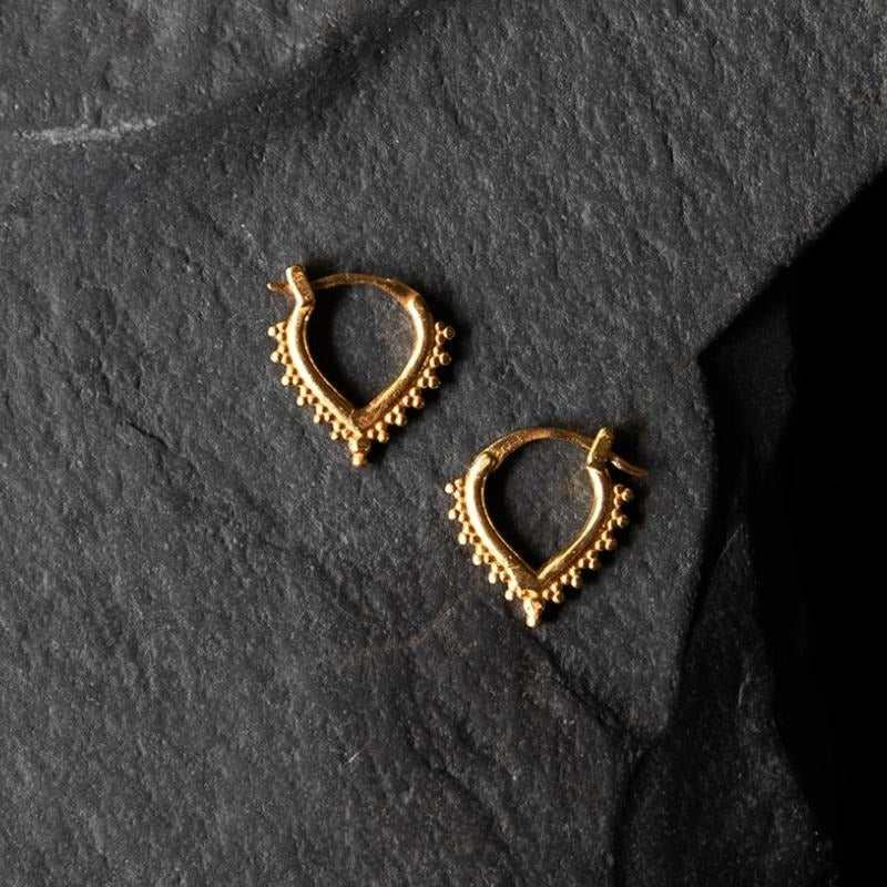 Paire de boucles d'oreilles en forme de goutte inversée dorée, ornées de petites perles délicates tout autour. Les anneaux présentent un design inspiré des bijoux traditionnels, offrant une touche élégante et intemporelle. Le fond en pierre noire texturée contraste magnifiquement avec la brillance du métal doré.