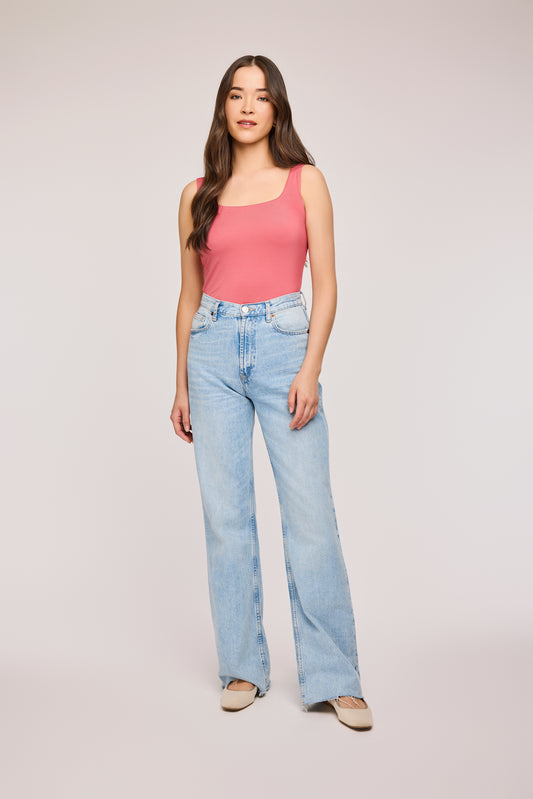 Débardeur à bretelles larges et encolure carrée. Le haut est d'un rose baie qui tire légèrement vers le rouge. Le modèle porte un jean taille haute bleu clair à jambes larges.Elle porte aussi des balerines beige. L'arrière-plan est un fond gris.