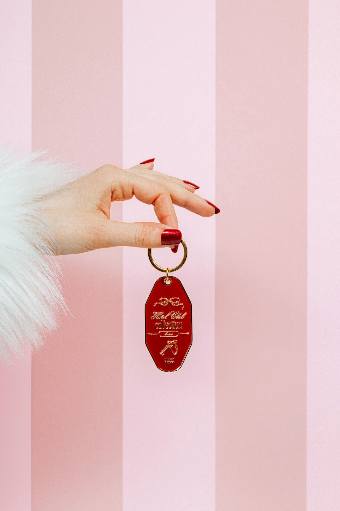 L'image montre une main élégante avec des ongles rouges vernis, tenant un porte-clés vintage rouge en forme de losange avec des inscriptions dorées. La main est partiellement couverte par une manche en fausse fourrure blanche. L'arrière-plan présente des rayures verticales roses et blanches, créant une ambiance rétro et sophistiquée.