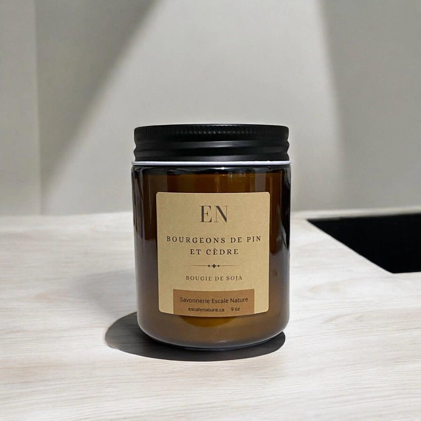 Bougie dans un pot en verre ambre avec un couvercle noir et une étiquette marron. En arrière-plan, on peut voir une table en bois et des décorations en rotin.