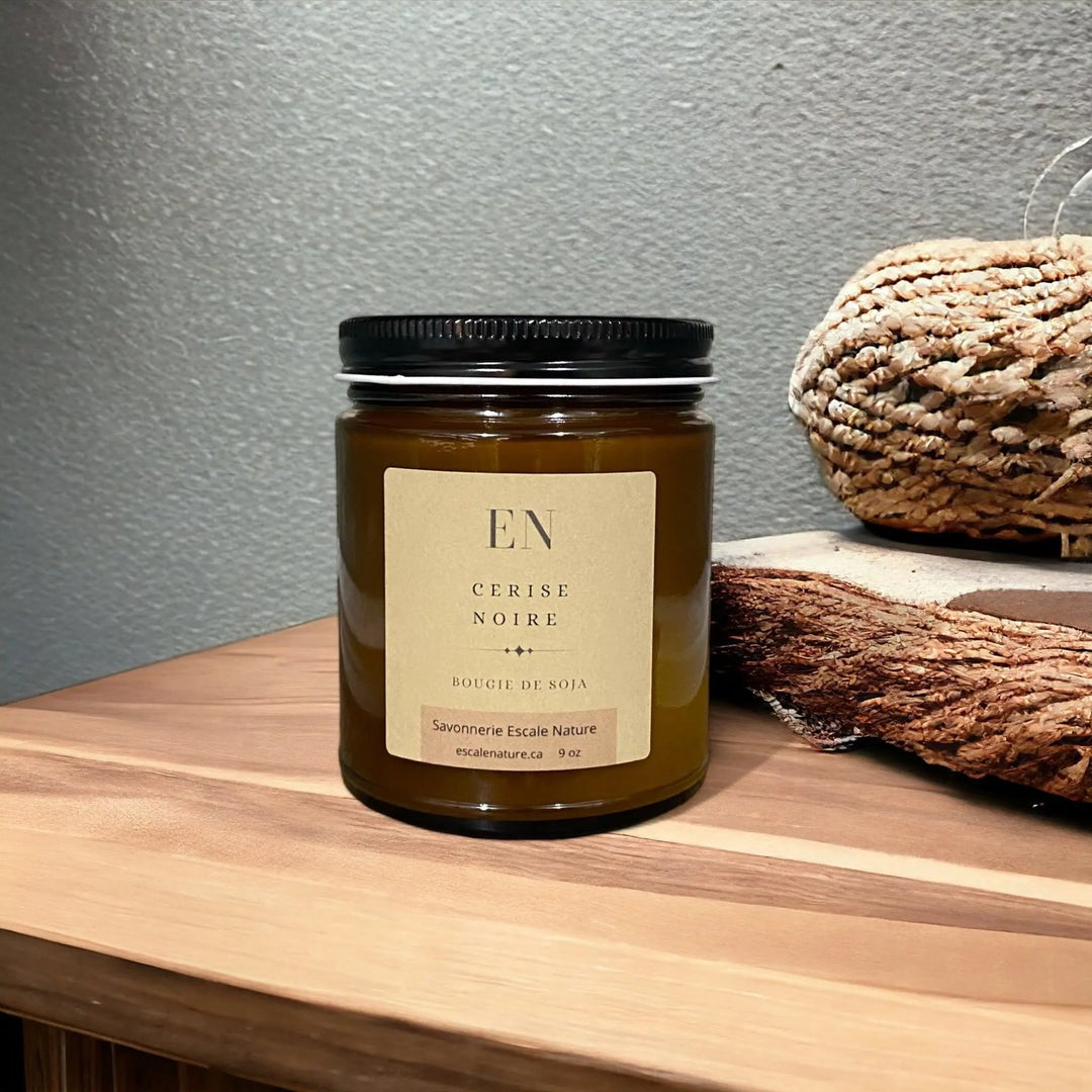 Bougie dans un pot en verre ambre avec un couvercle noir et une étiquette marron.En arrière-plan, on peut voir une table en bois et des décorations en rotin.