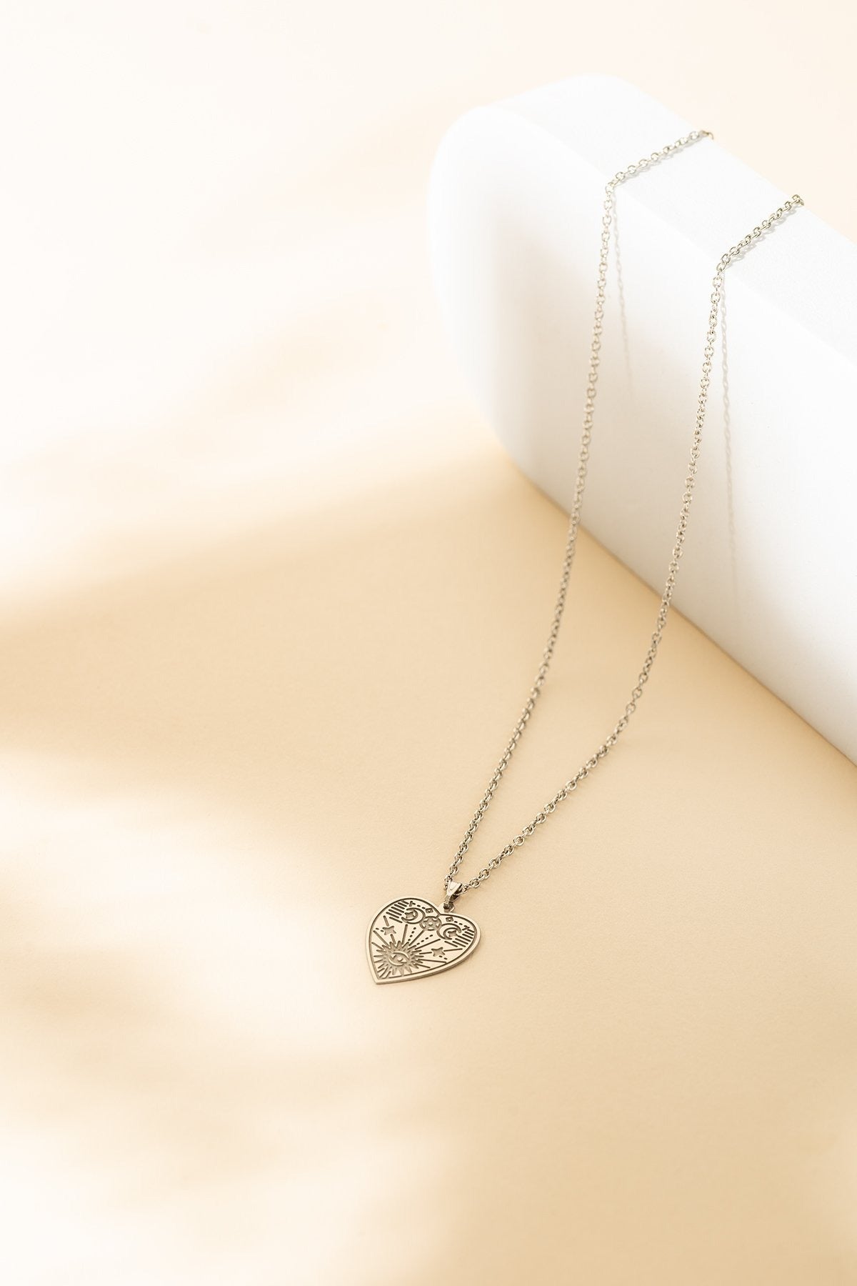 Collier minimaliste avec breloque en forme de coeur avec gravures. On peut y voir, un oeil, un soleil, des étoiles et une lune. Le collier est posé sur une pièce en céramique blanche et sur un fond beige.