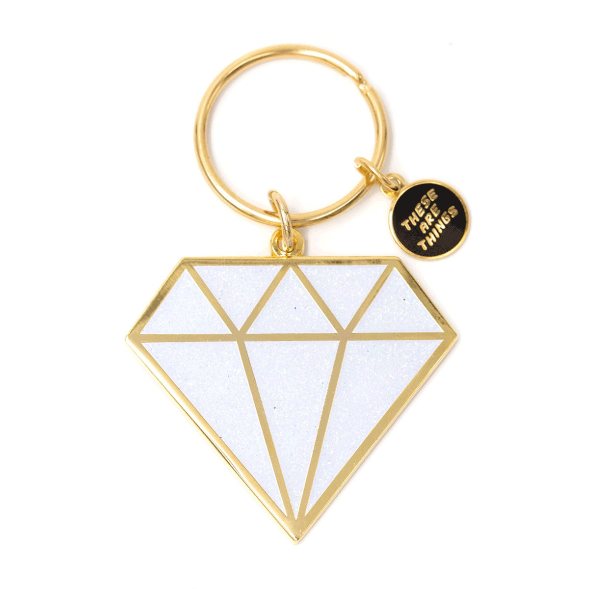 Porte-clef et breloque en forme de diamant. Le porte-clef est en émail de couleur or. Le diamant est blanc et pailleté. Il est découpé par des lignes or. Il y a une toute petite breloque sur laquelle est inscrit le nom de la marque qui est There are things. L’arrière-plan est un fond blanc.