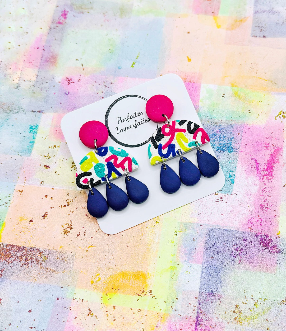 La partie collée à l'oreille est un rond fushia sur lequel est attaché un demi-cercle blanc recouvert de lignes de plusieurs couleurs. Il y a trois gouttes bleu foncées suspendues à chaque boucle d'oreille. Les boucles d'oreilles sont présentées sur un carton blanc avec le logo de Parfaites Imparfaites. L'arrière-plan est multicolore.