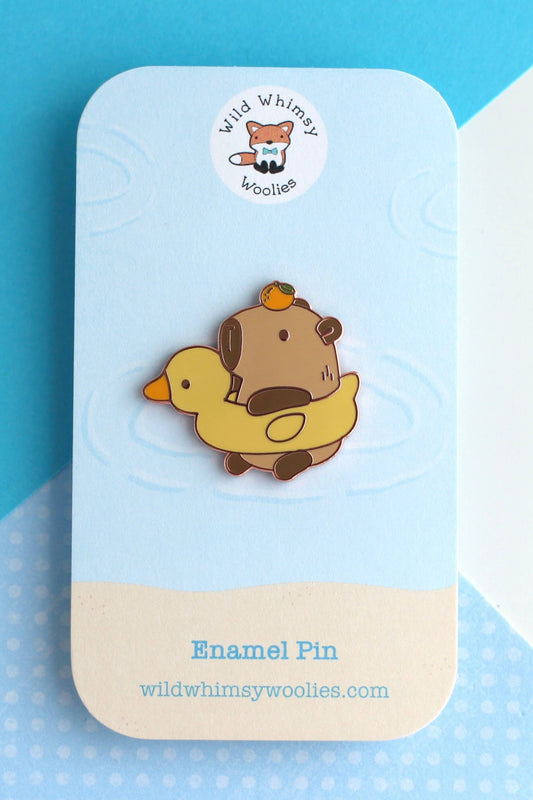 Épinglette Capybara et Canard en émail dur cuivré, présentée sur carte bleue avec logo Wild Whimsy Woolies.