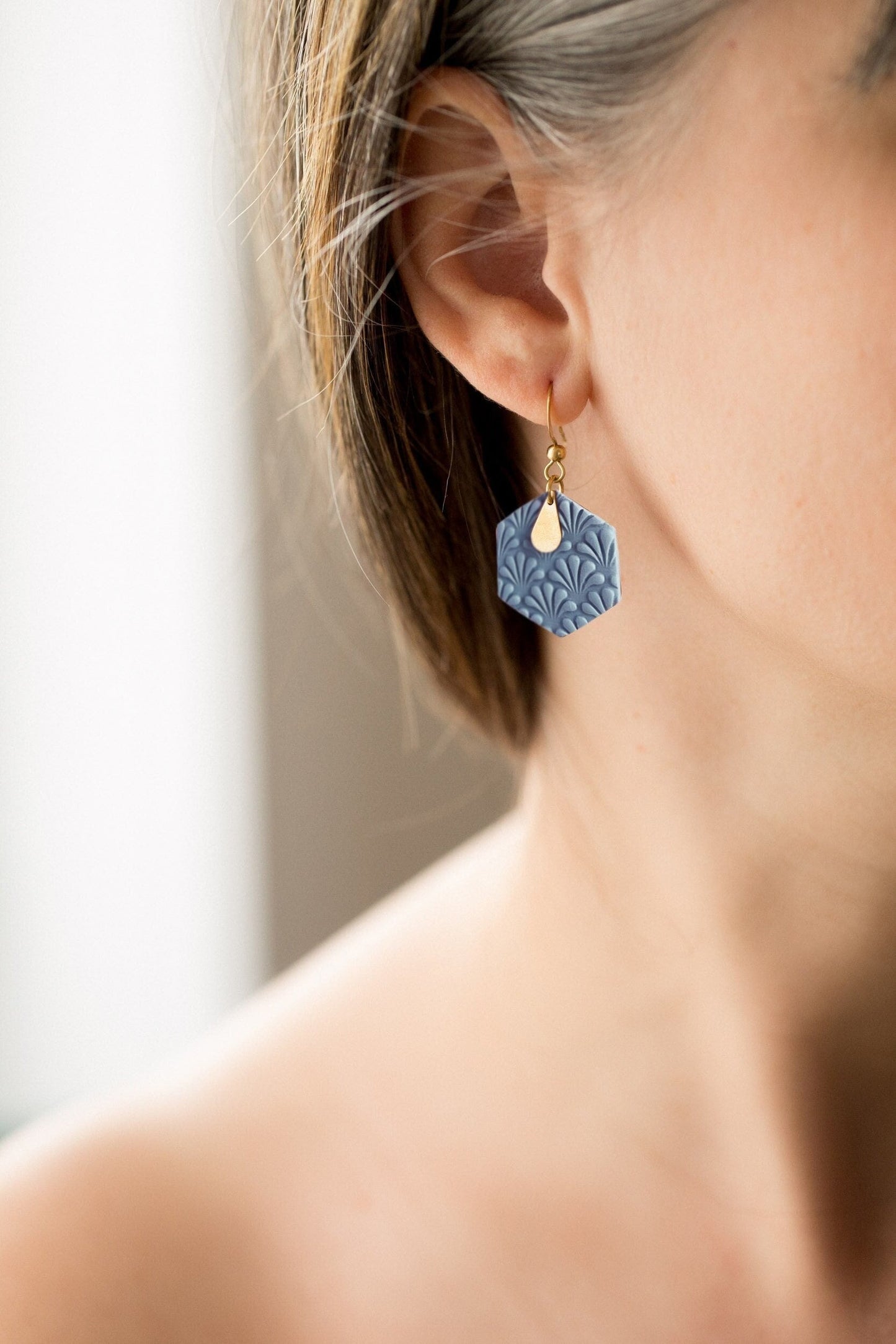 Boucles d'oreilles en argile polymère bleu en forme d'hexagone avec une petite plaque en laiton en forme de goute. Le modèle se tient de profil, c'est un plan rapproché.