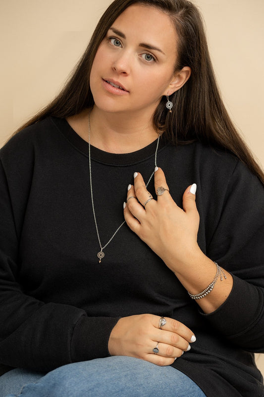 Sur cette photo, on peut voir une personne assise. Elle a une main sur sa cuisse et l'autre main près de son visage. Elle porte un pull noir, un jean, le collier Fjolla et d'autres bijoux de la marque. L'arrière-plan est un fond beige.