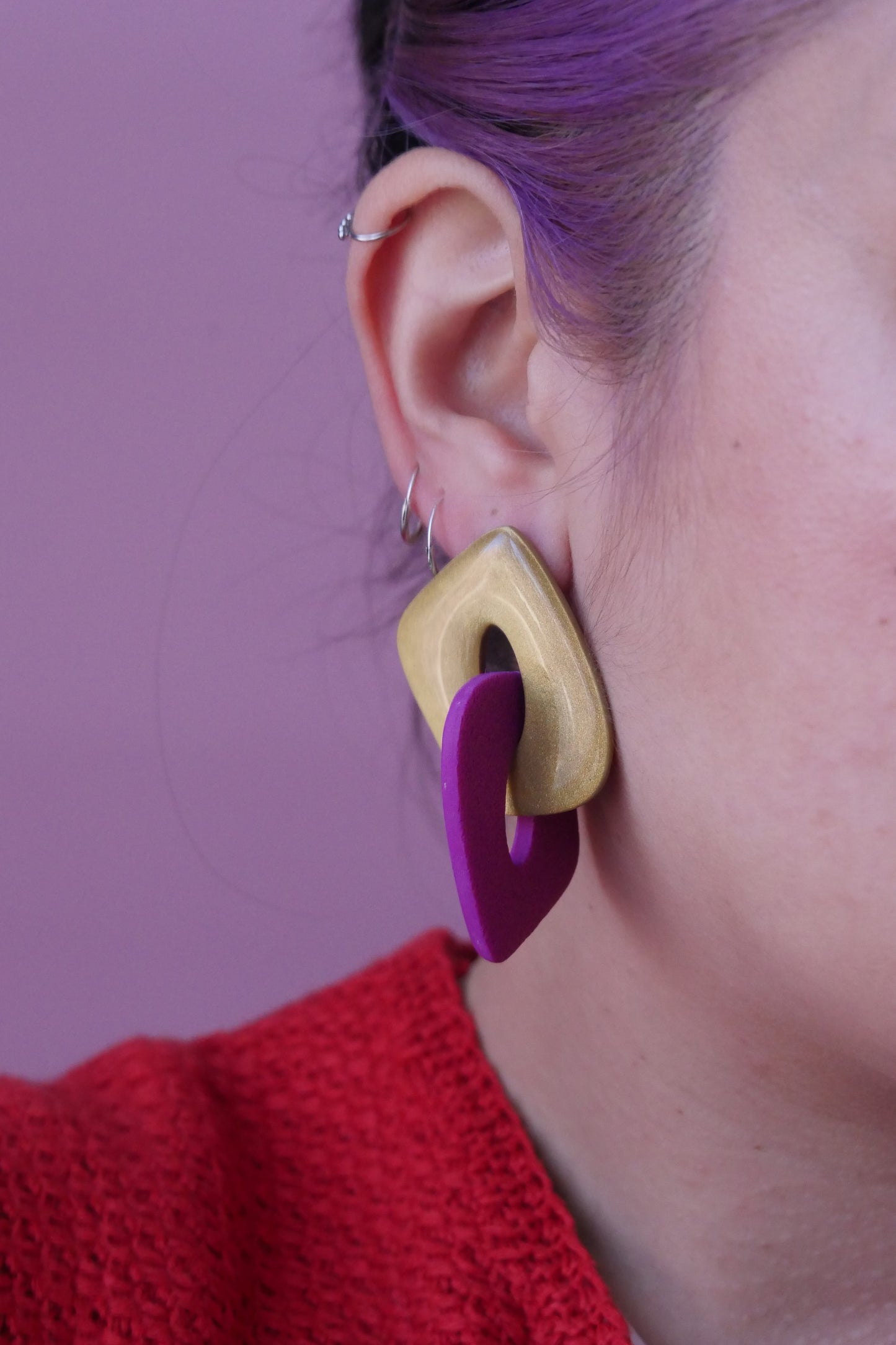 Les boucles d'oreilles sont faites de deux triangles creux et arrondies qui s'entremêlent. Un est à paillettes dorées et l'autre fushia. Plan rapproché sur l'oreille d'un modèle portant une boucle d'oreilles et trois petits anneaux fins argent.