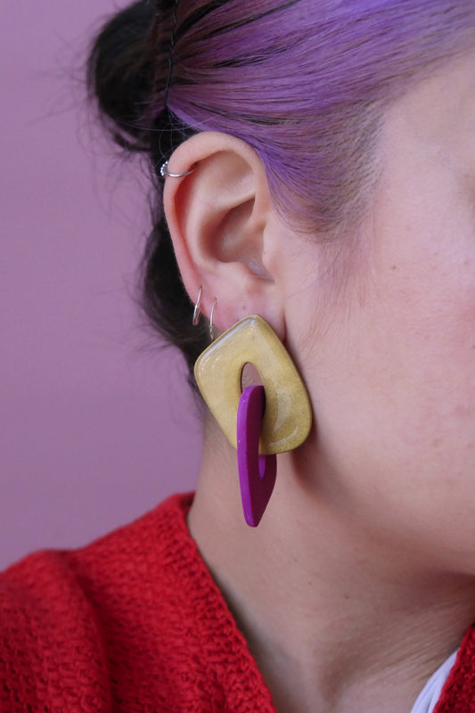 Les boucles d'oreilles sont faites de deux triangles creux et arrondies qui s'entremêlent. Un est à paillettes argentées et l'autre fushia. Plan rapproché sur l'oreille d'un modèle portant une boucle d'oreilles et trois petits anneaux fins argent.