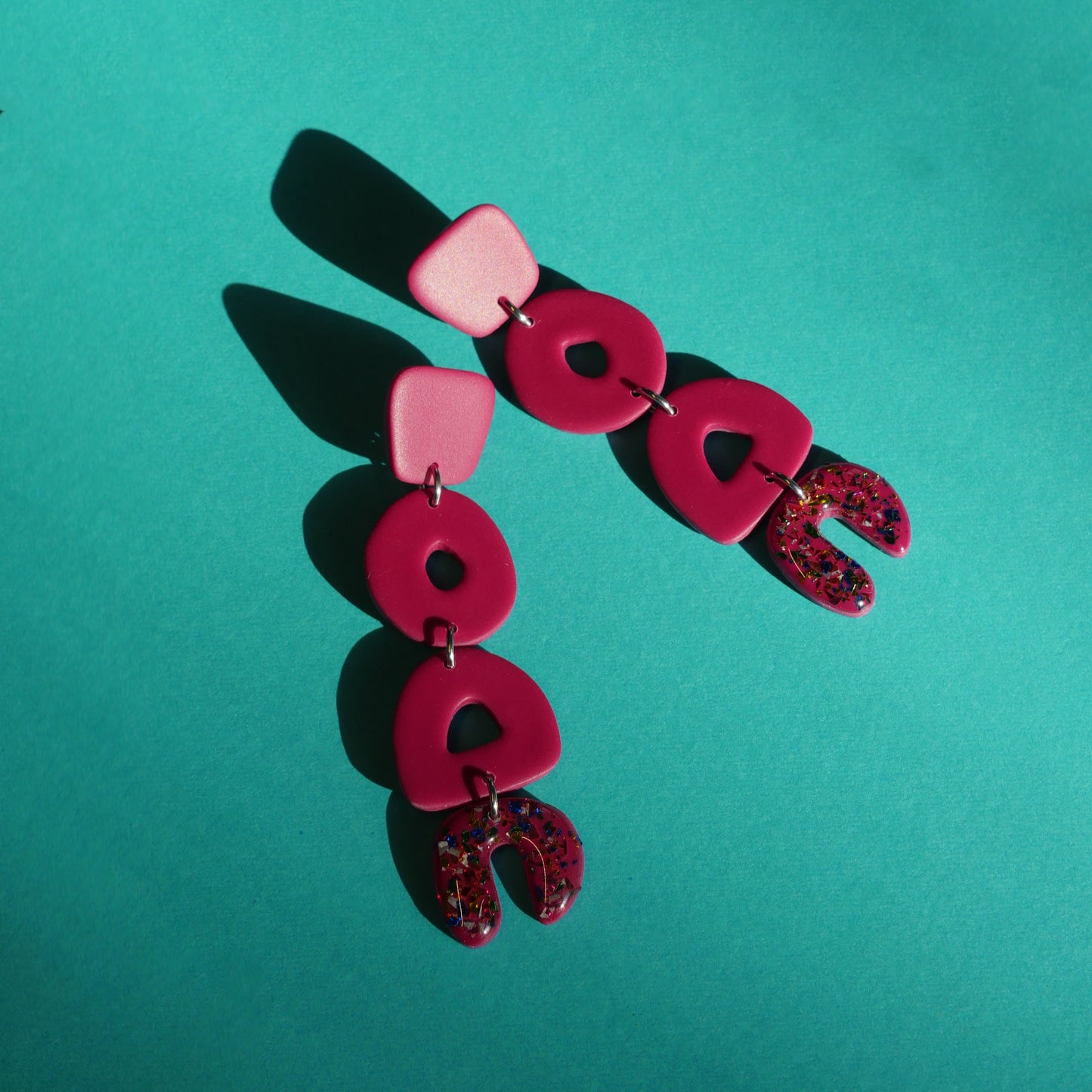 Longues boucles d'oreilles faites de quatres pièces de formes différentes et abstraites. Il y a des paillettes sur la dernière pièce. L'arrière-plan est un fond vert.
