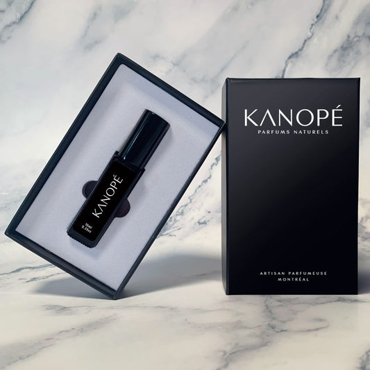 Ici, on peut voir que le parfum vient dans une boite en carton noir. Sur le couvercle, en haut on peut lire Kanopé et en de-dessous parfums naturels. En bas, il est écrit: artisan parfumeuse, Montréal. L'intérieur de la boîte est blanc. Sur la bouteille, on a collé une étiquette noire sur laquelle on peut lire Kanopé et le volume de la bouteille qui est de 10 ml ou 0,33 oz. L'arrière-plan est un fond en marbre gris.