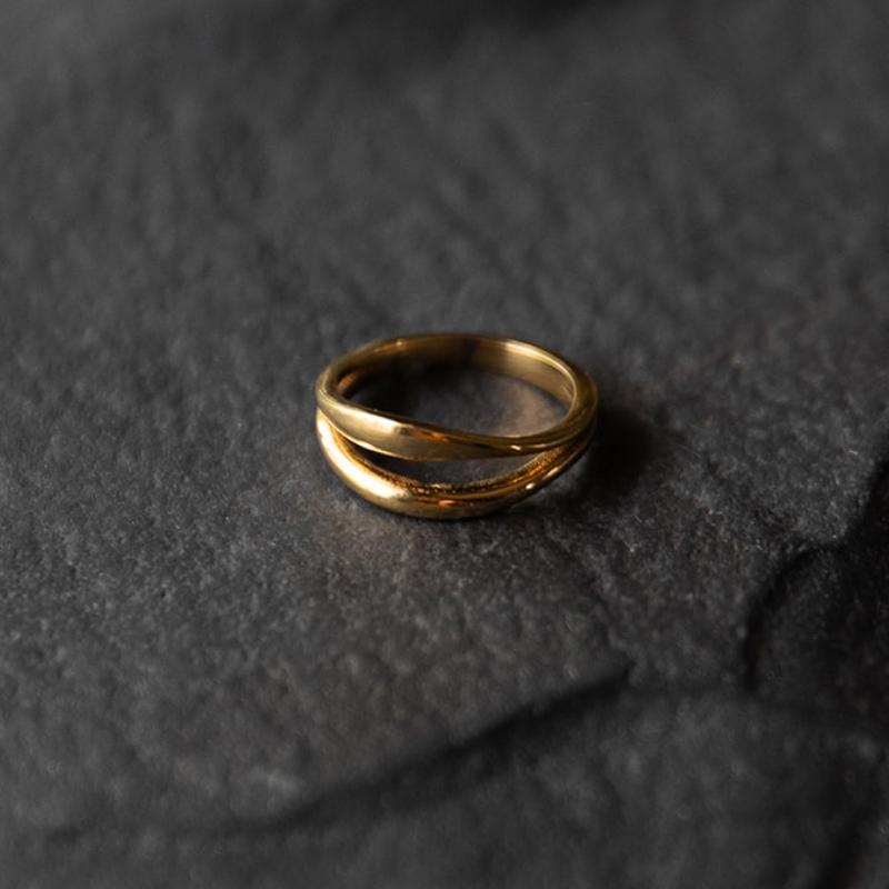 Bague dorée minimaliste avec un design entrecroisé. Les deux bandes fines se croisent délicatement, créant un effet de mouvement fluide et élégant. Le fond en pierre noire texturée met en valeur la finition brillante et raffinée du métal doré.