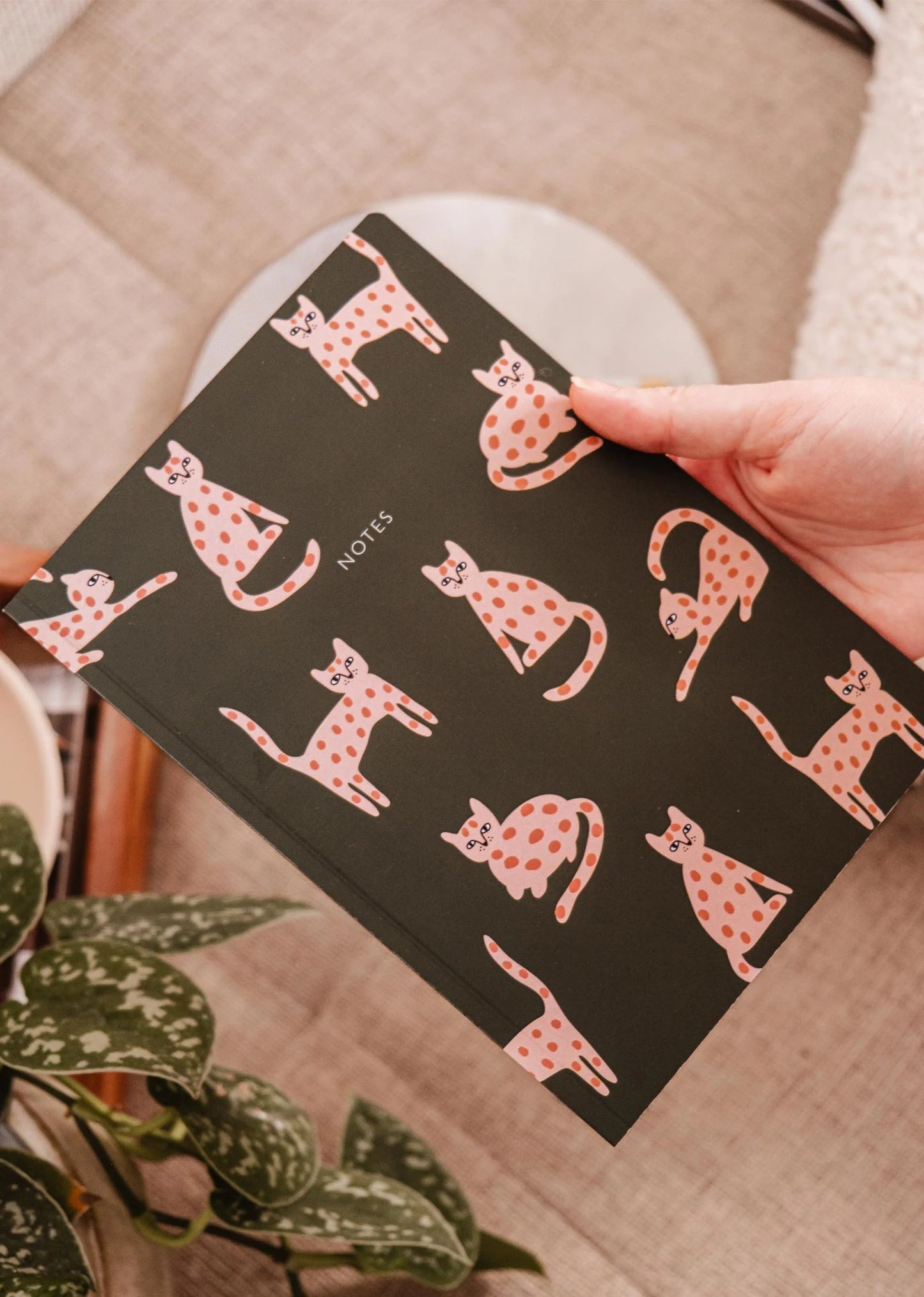 La couverture du carnet de notes est verte foncé avec des petits chats rose tachetés dans plein de positions rigolotes. Quelqu'un tient le carnet dans sa main. En arrière-plan, on voit une plante.