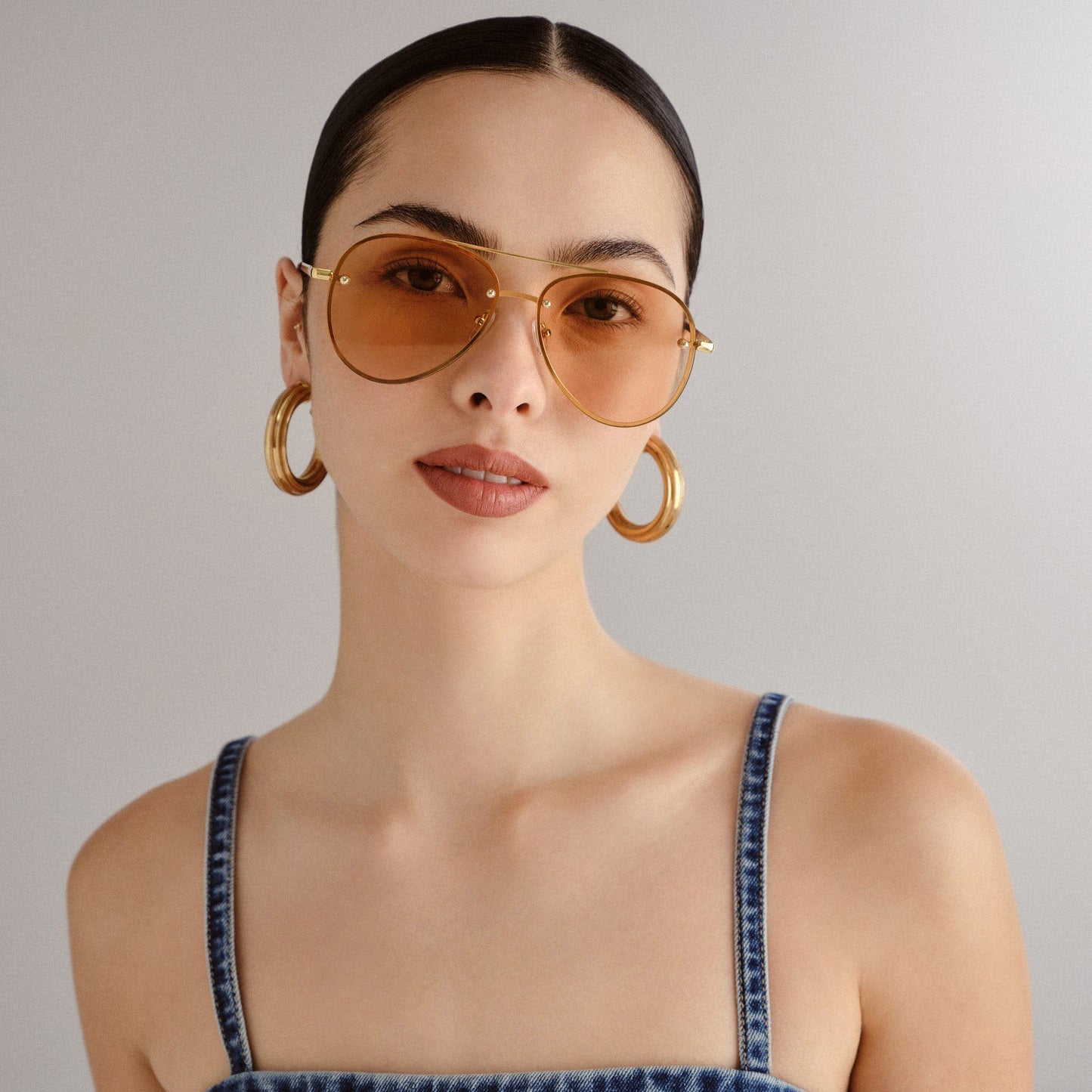 Ces lunettes de soleil ont une monture en métal doré, fine et légère, avec un design aviateur intemporel. Les verres sont teintés dans une nuance ambrée, apportant une touche chaleureuse et rétro tout en protégeant les yeux de la lumière. La barre supérieure de la monture traverse horizontalement le haut des lunettes, renforçant leur style classique. Les branches sont également en métal doré, avec des embouts délicats pour un confort optimal. Ces lunettes sont portées par une femme avec des cheveux lissé.