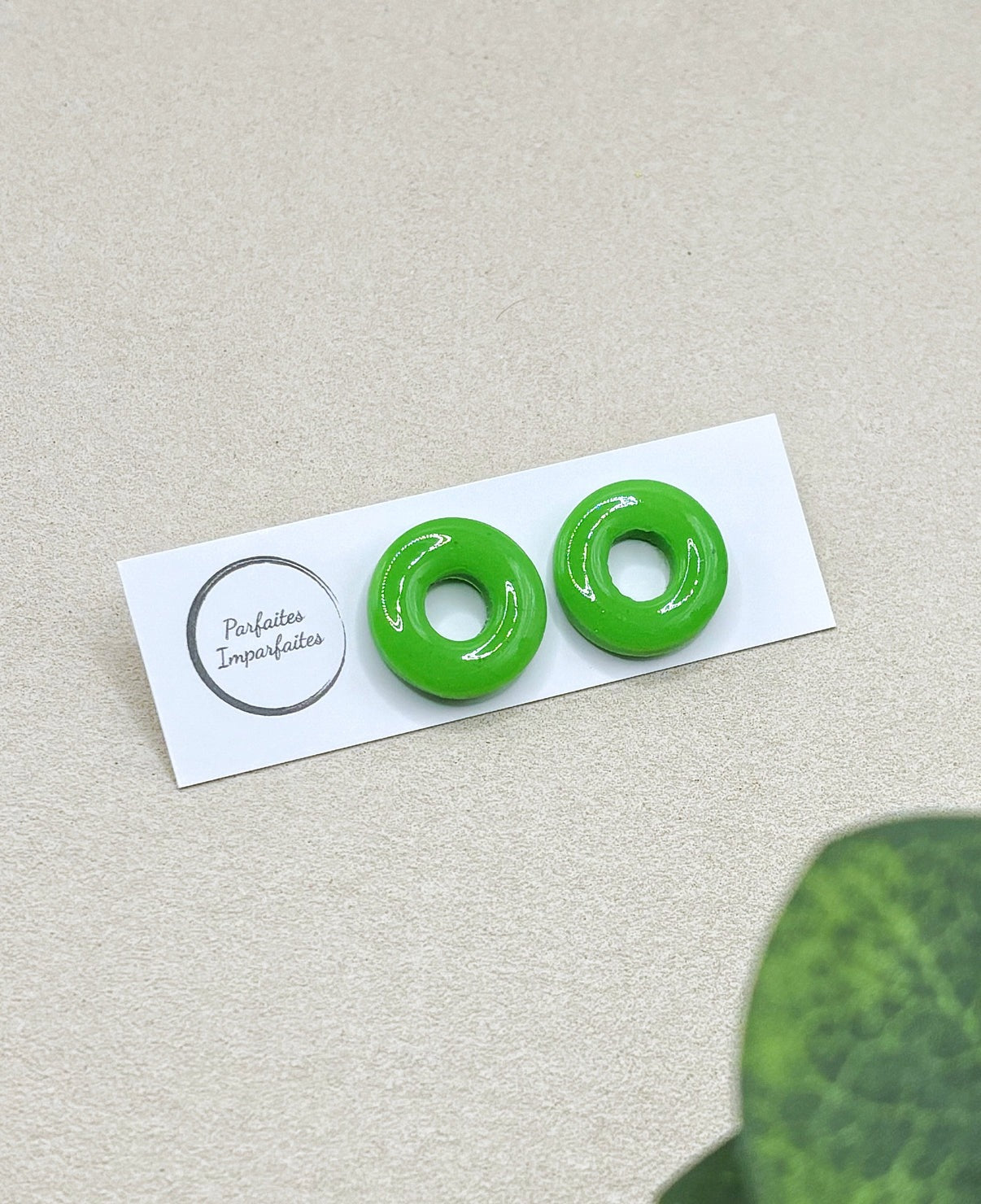 Petites boucles d'oreilles ronde avec un trou au centre. Elles sont vernis ce qui leur donne vraiment l'apparence de bonbons. Elles sont entièrement vertes pomme. Les boucles d'oreilles sont présentées sur un carton blanc avec le logo de la marque Parfaites Imparfaites et l'arrière-plan est un fond beige.