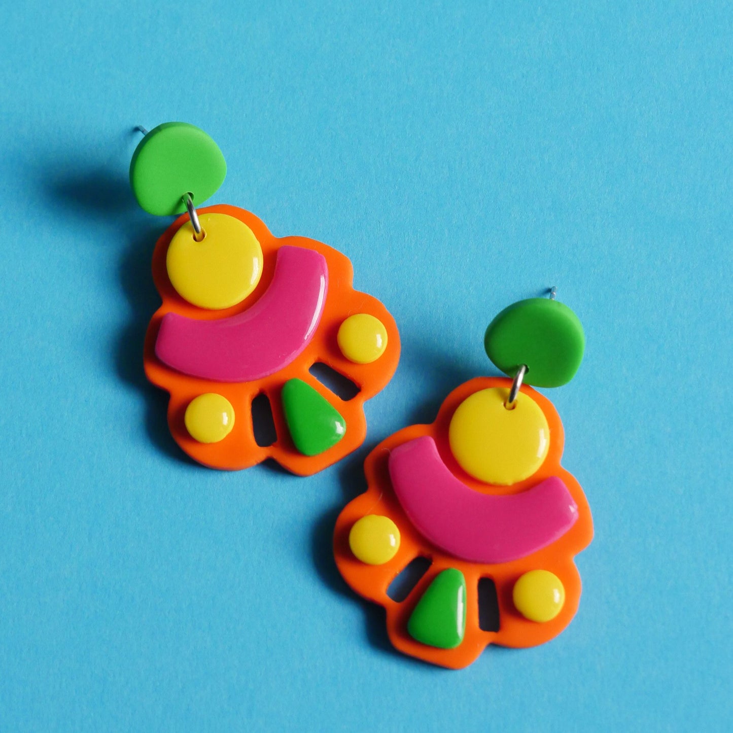 Boucles d'oreilles en forme abstraite, plates de couleur fluo (orange, rose, vert et jaune). L'arrière-plan est un fond bleu clair.