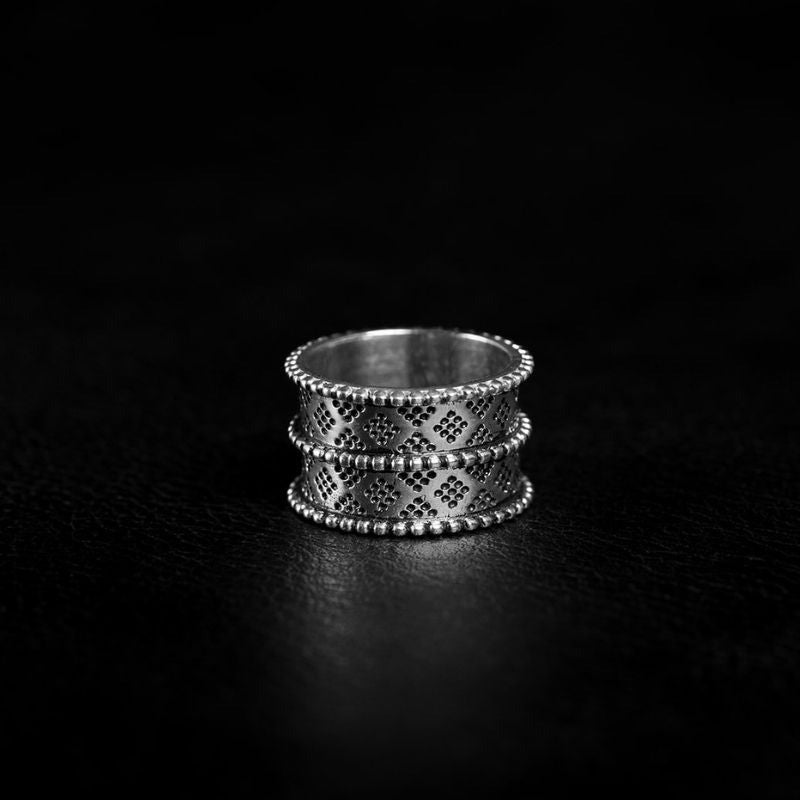 Bague en argent au design large et ornemental, avec des motifs géométriques en losanges finement gravés. Les bords de la bague sont décorés de petites perles métalliques, ajoutant une touche élégante et texturée. Le fond noir profond met en valeur la brillance argentée et les détails sophistiqués de ce bijou intemporel.