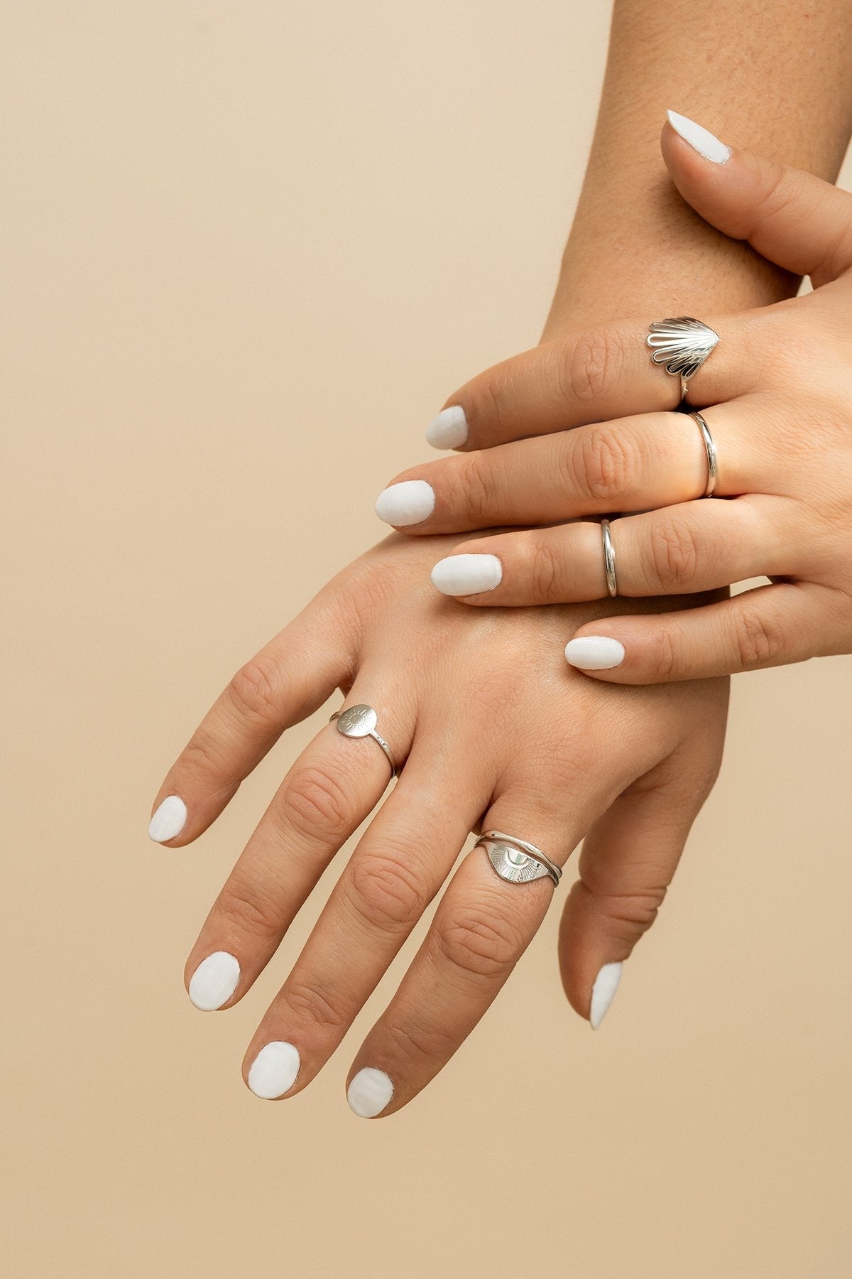 Sur cette photo, on peut voir les mains d'une même personne qui se croisent. Elle porte la bague Piki en argent et d'autres bagues de la même marque. Elle a une manucure blanche. L'arrière-plan est beige.