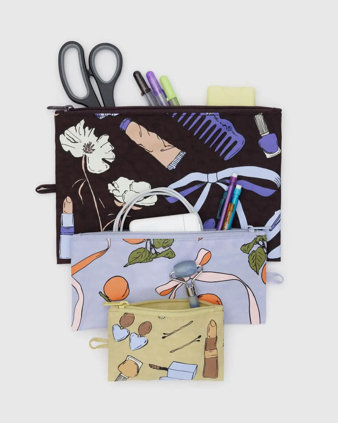 Ensemble de trois pochettes plates en tissu, de tailles différentes et aux imprimés élégants. La plus petite est jaune pâle avec des illustrations de maquillage, d'épingles à cheveux et de boucles d'oreilles. La moyenne est bleu lavande avec un motif d'abricots et de rubans. La plus grande est marron foncé avec des illustrations de fleurs, de peignes et d'accessoires de beauté. Chaque pochette possède une fermeture éclair sur le dessus et une petite languette latérale pour une ouverture facile.