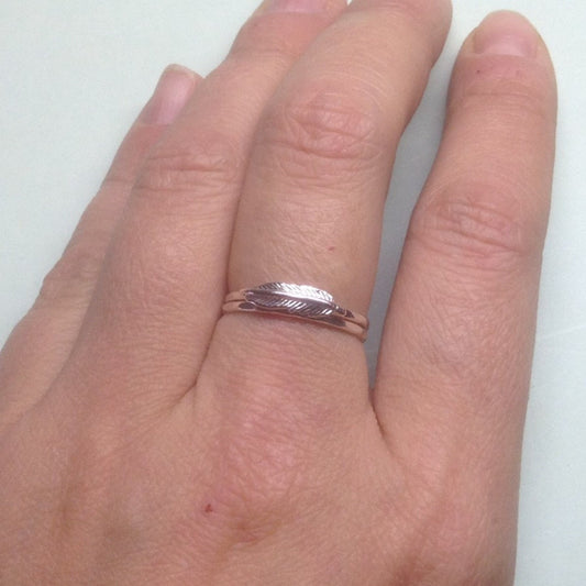 Bague délicate sur laquelle est soudée un petite plume en argent sterling. L’anneau est une fine bande en argent martelé. La bague est porté au majeur gauche d’une personne. Elle est superposé sur un anneau identique à la bague, mais sans breloque.