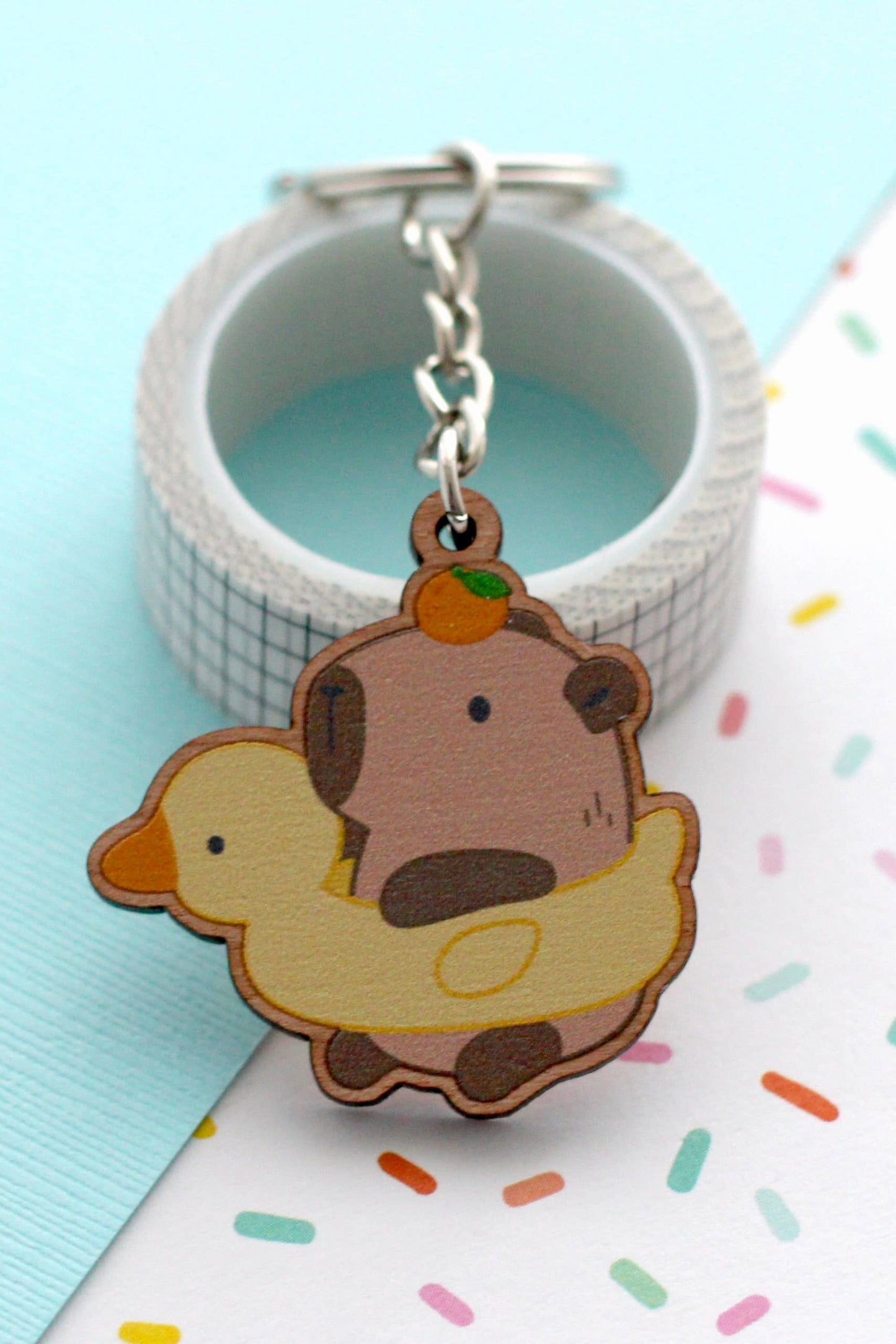 Porte-clés en bois représentant un capybara avec une bouée canard et une orange sur la tête, posé sur fond pastel turquoise et blanc.