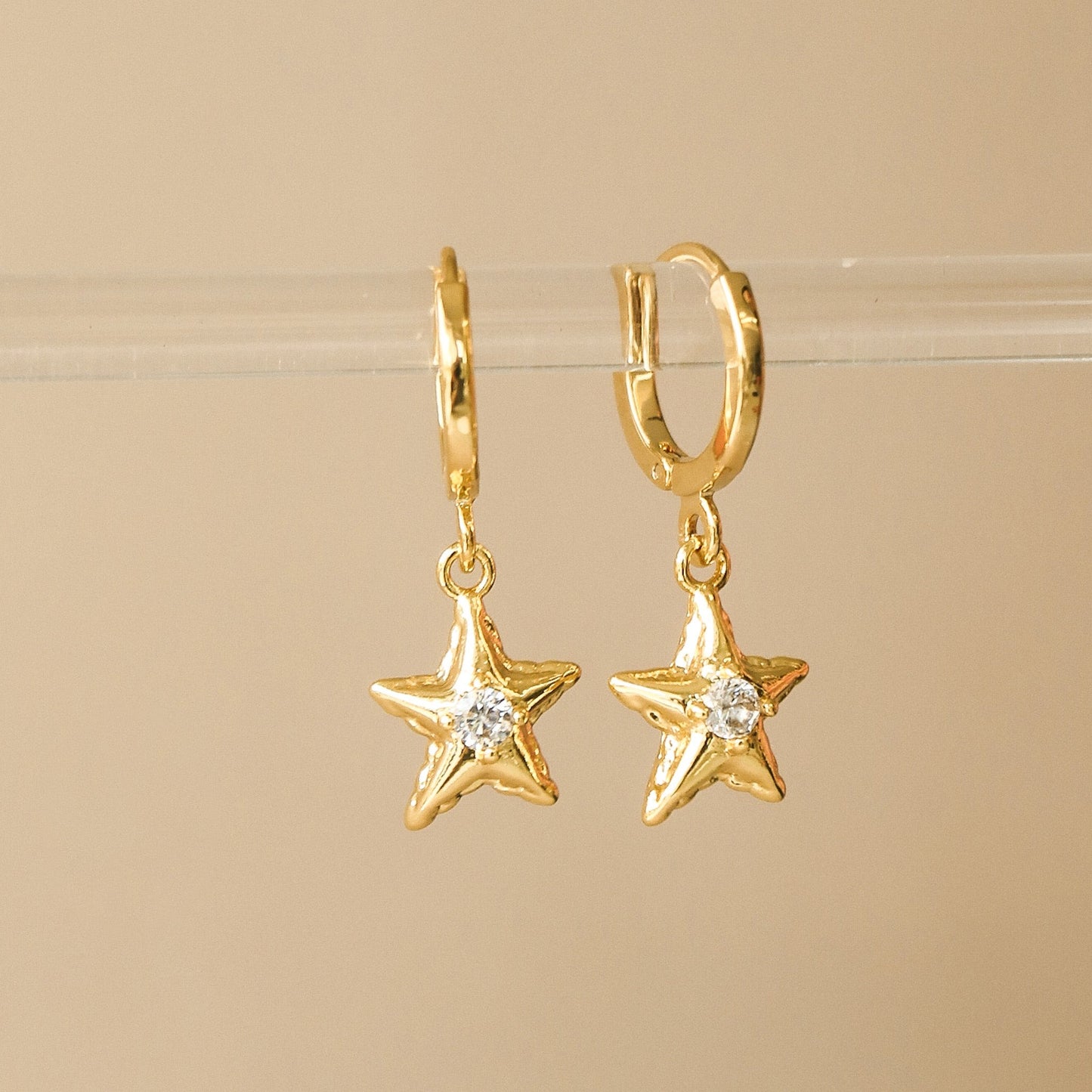 Anneau en laiton plaqué or avec un pendentif en forme d'étoile de mer avec un zircon en son centre. Les boucles d'oreilles sont accrochées sur un présentoir en acrylique transparent sur sur un fond beige.