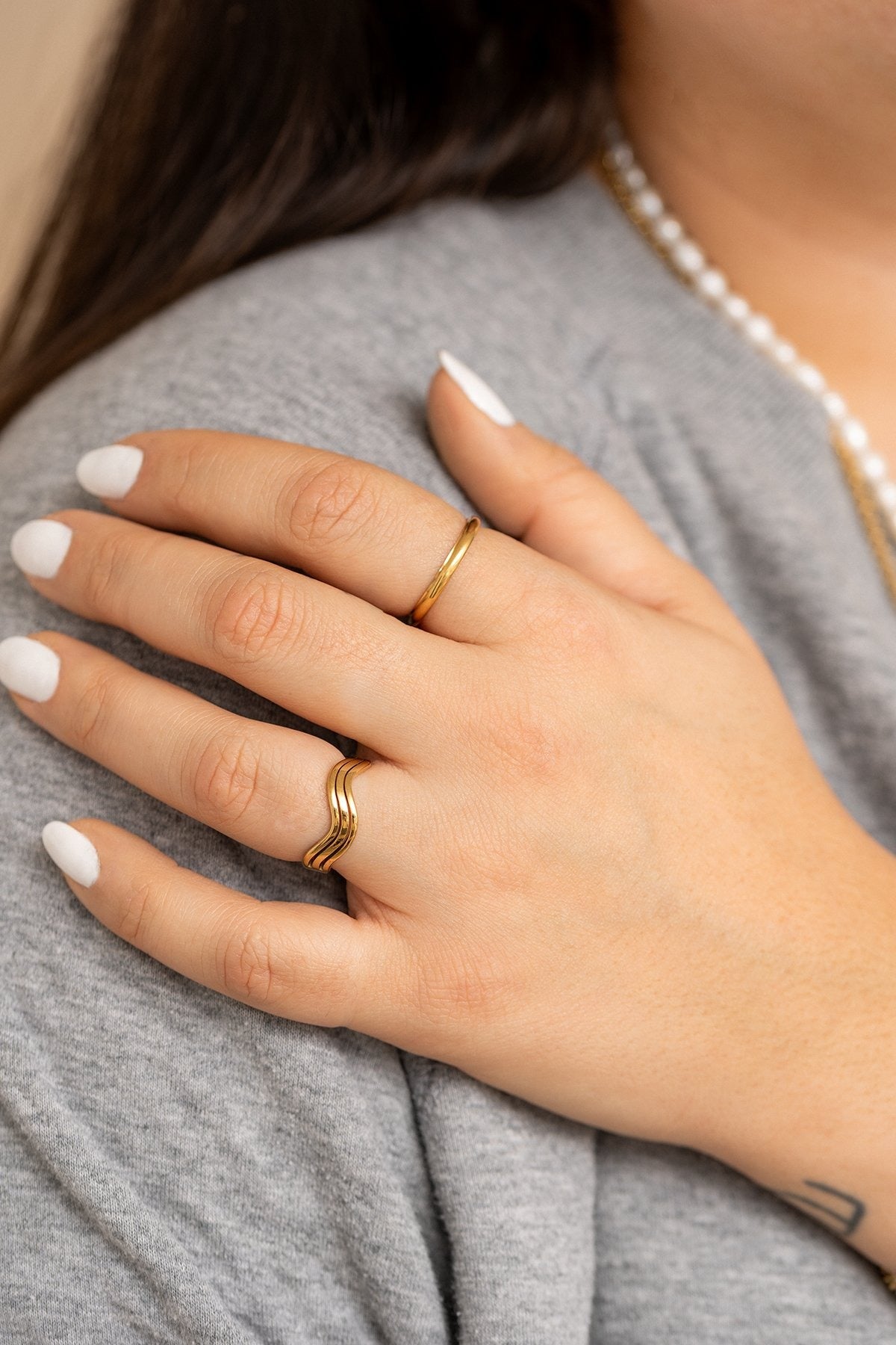 Ici on voit un plan rapproché sur une personne. Sa main est posé sur son épaule. Elle porte un t-shirt gris. Elle a une manucure blanche. Sur sa main, on voir la bague wavy à son annulaire et une bague très fine sur l'index.