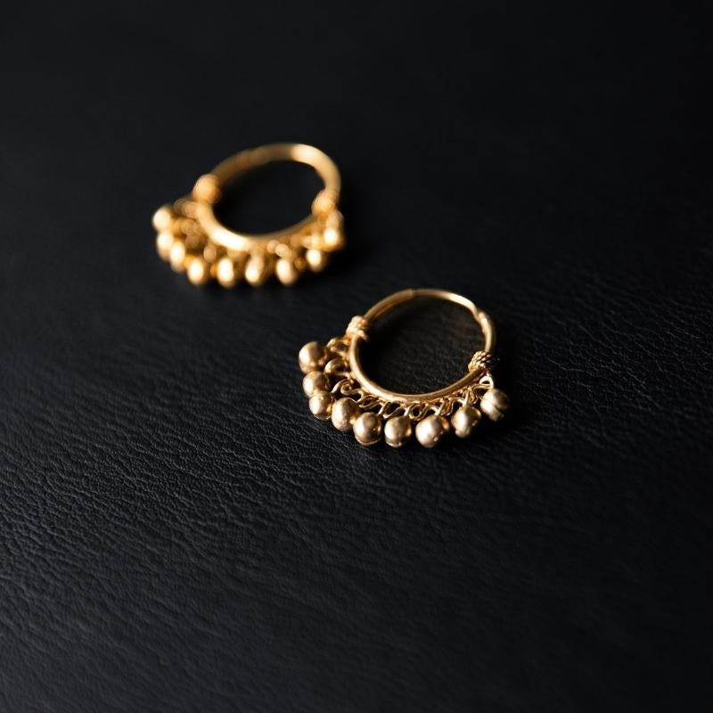 Paire de boucles d'oreilles dorées en forme de petits anneaux ornés de perles métalliques. Chaque anneau présente une rangée de petites perles dorées suspendues, offrant un mouvement délicat et un charme bohème. Le fond noir texturé fait ressortir la brillance et le design raffiné de ces boucles d'oreilles.