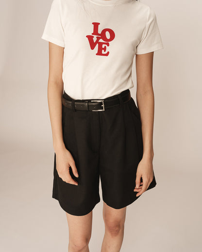 Bermuda noir élégant à taille haute avec une coupe fluide, porté par un mannequin avec un t-shirt blanc et des escarpins noirs.