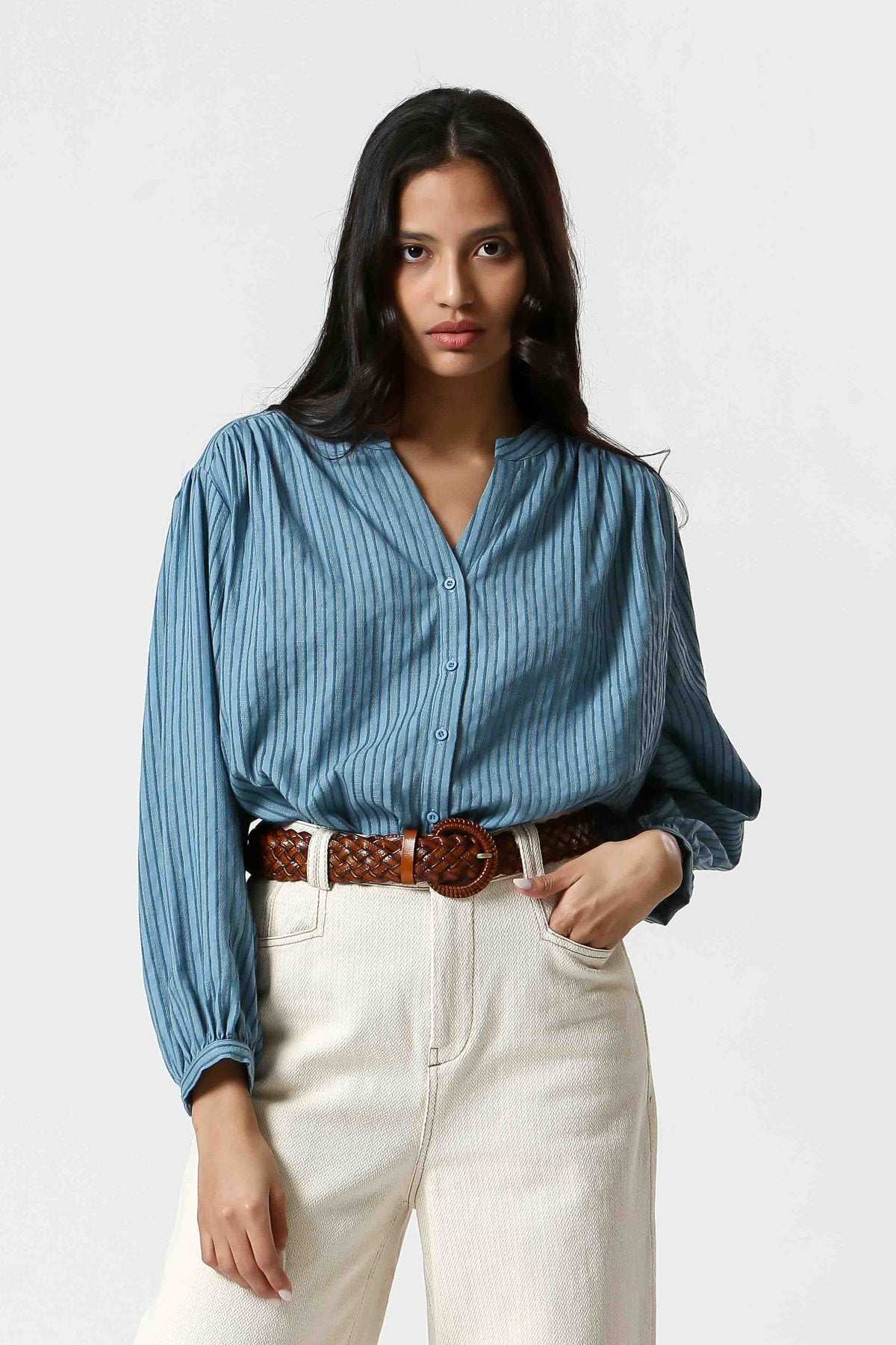 Blouse bleu clair en coton et lin, avec boutonnière devant, effet ligné texturé en broderie, manches légèrement bouffantes, décolleté en V et poignets resserrés.