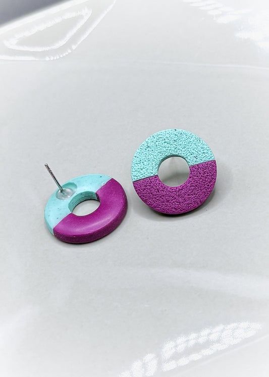 Petites boucles d'oreilles rondes avec un trou au milieu. Elles sont texturées avec un papier ponce qui leur donne un aspect de pierre. Elles sont divisées en deux couleurs, la partie en haut est turquoise et celle d'en bas mauve. Une des boucles d'oreilles est à l'envers, on peut voir que le derrière est vernis. Les boucles d'oreilles sont présentées sur une assiette blanche. 