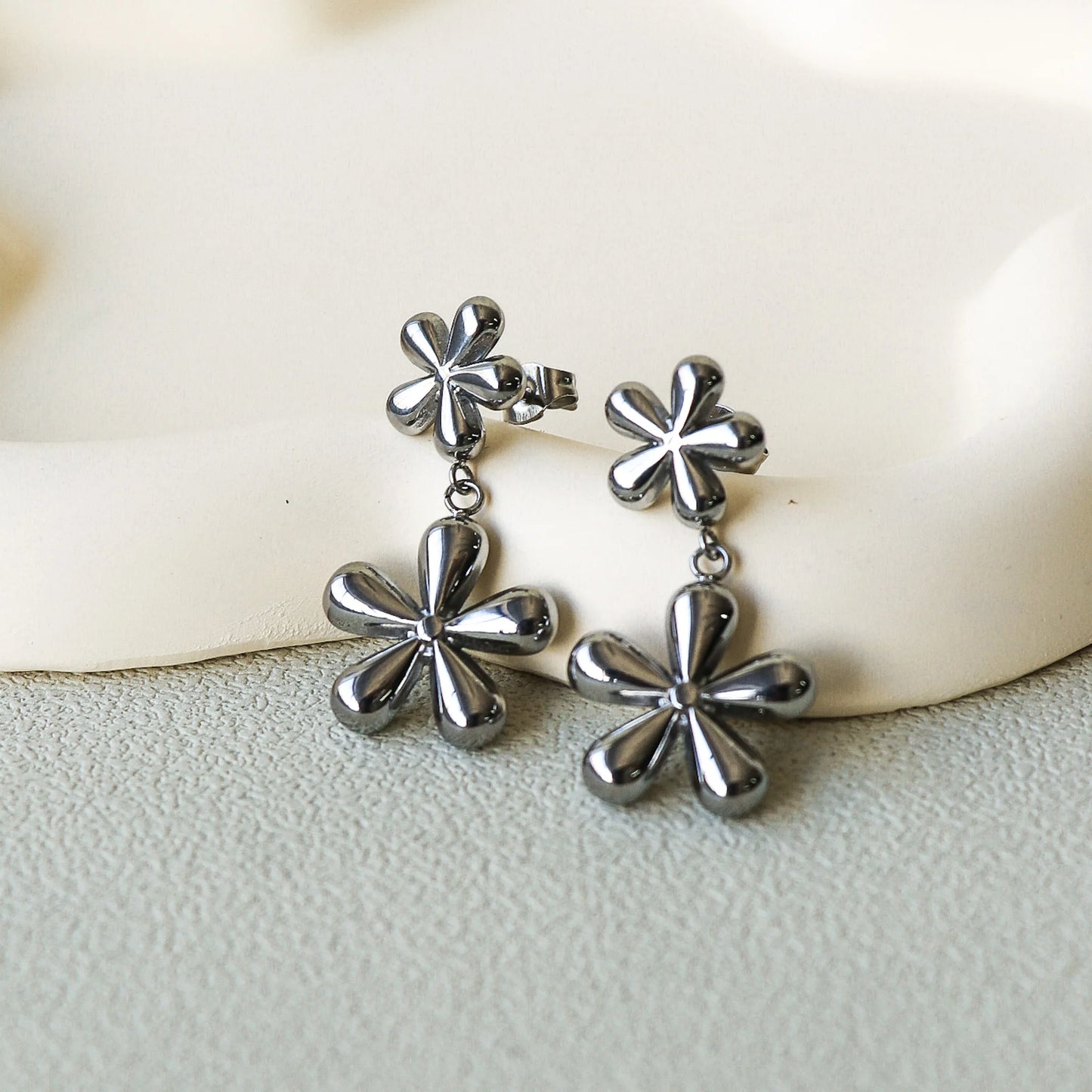 Boucles d'oreilles en forme de fleurs, ce sont deux fleurs de tailles différentes suspendue l'une à l'autre. La paire est faite en acier inoxydable couleur argent. Les boucles d'oreilles sont déposées sur une assiette en céramique beige sur une table grise. Plan rapproché sur les boucles d'oreilles. 