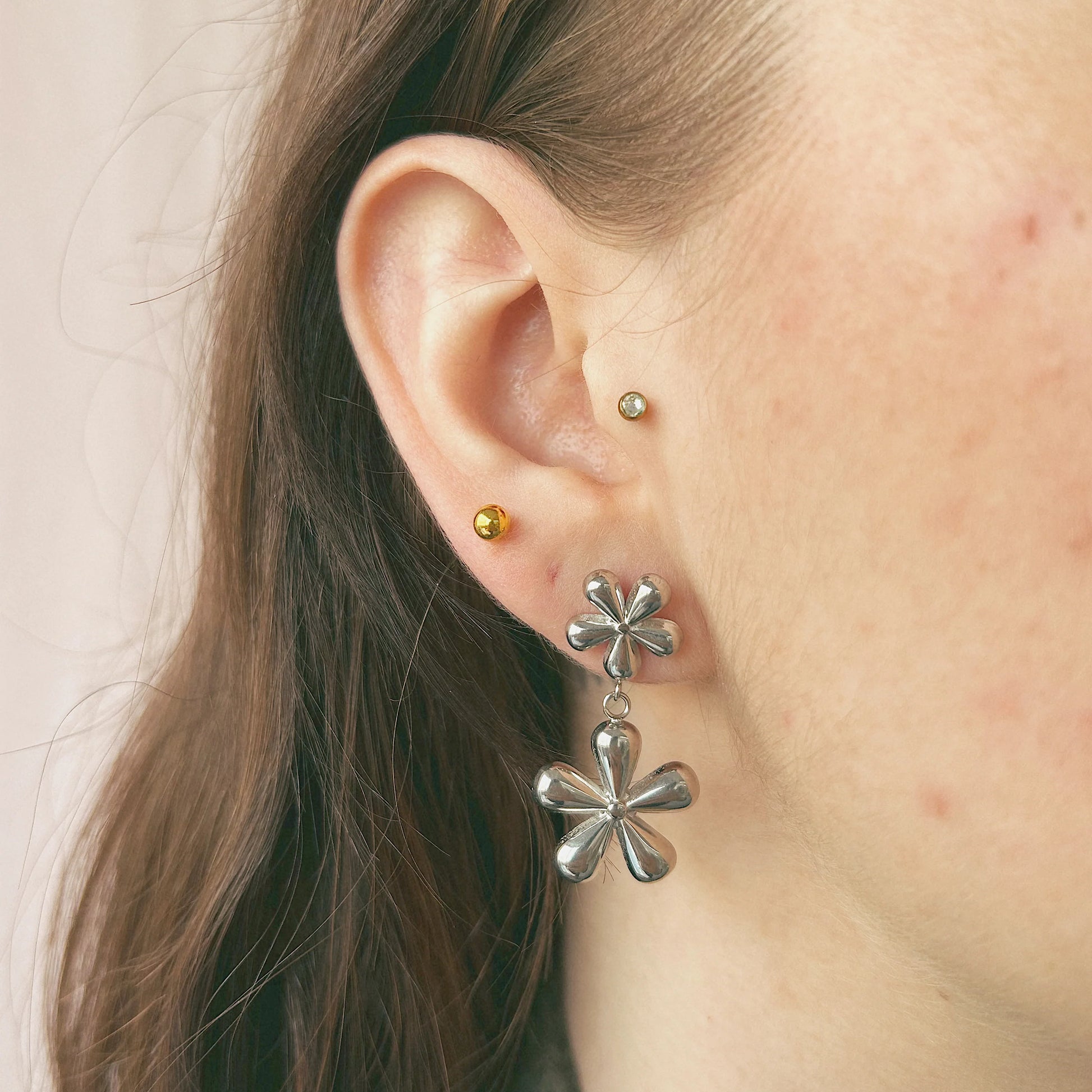 Boucles d'oreilles en forme de fleurs, ce sont deux fleurs de tailles différentes suspendue l'une à l'autre. Un modèle de profil porte la paire de boucles d'oreilles ainsi qu'un petit clou d'oreille doré de la même marque. 