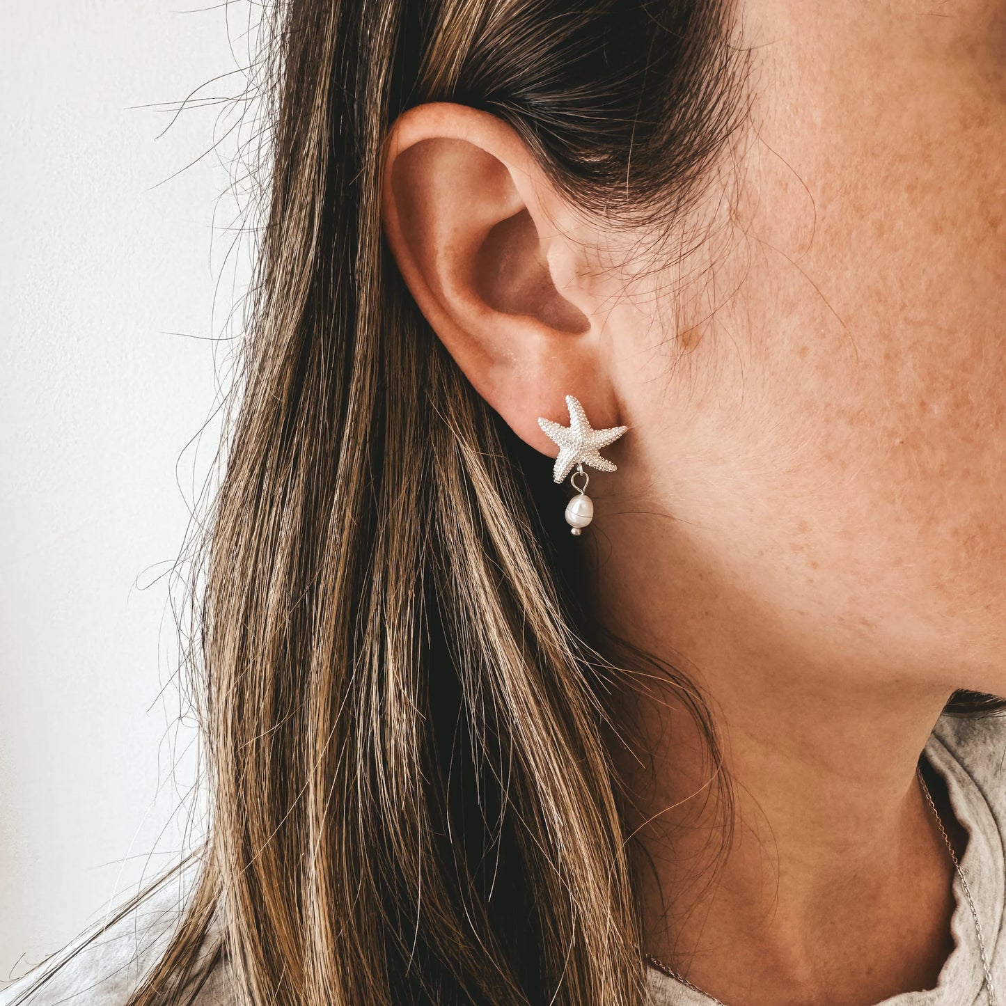 Boucles d'oreilles en forme d'étoiles de mer texturées avec perles d'eau douce. Elles sont faites en laiton plaqué argent clair. Le modèle se tient de profile est porte les boucles d'oreilles Demeria.