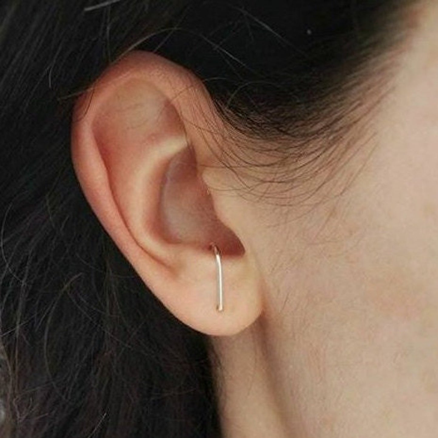 Petite boucles d'oreille en forme de J fabriquée en argent 925. Le creux du J vient s’accrocher dans l’intérieur de l’oreille au-dessus du lobe. La boucle d’oreille est porté par une personne, on voit son oreille au complet.  