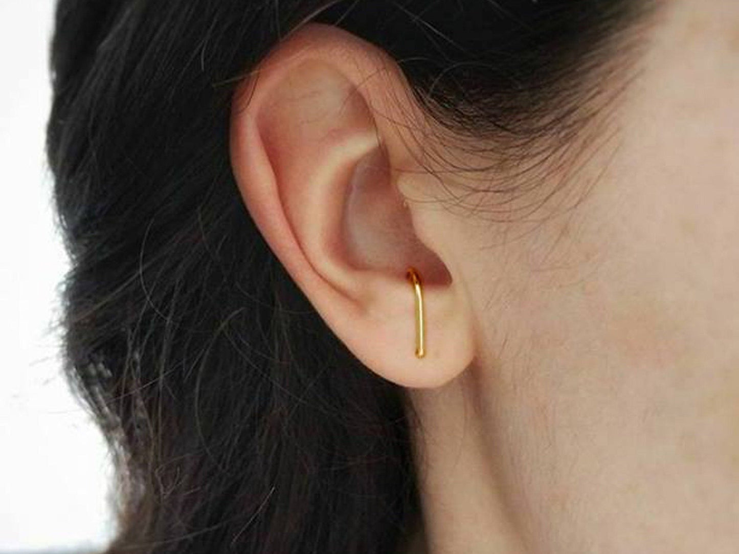 Petite boucles d'oreille en forme de J fabriquée en or. Le creux du J vient s’accrocher dans l’intérieur de l’oreille au-dessus du lobe. La boucle d’oreille est porté par une personne, on voit son oreille au complet.  