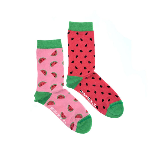 Paire de chaussettes dépareillées sur fond blanc. Pour les deux chaussettes, les extrémités et les talons sont en vert. Sur une chaussette on peut voir des tranches de pastèques sur un fond rose et sur l’autre chaussette des pépins sur un fond rouge.