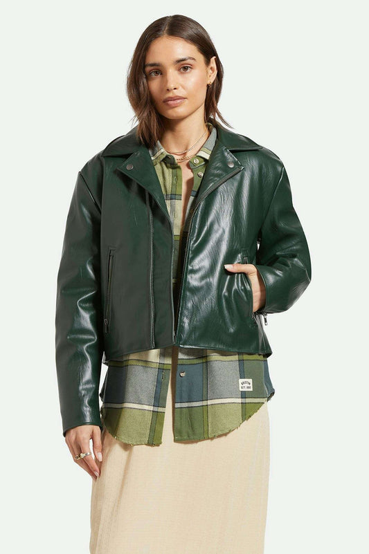 Manteau type perfecto en simili cuir lisse vert forêt porté par une femme. Le manteau est superposé sur une chemise et une robe longue. Vu de devant.