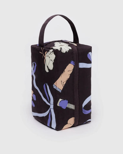 Trousse de toilette rectangulaire en tissu noir avec un motif illustré de rubans violets, fleurs et accessoires de beauté. Elle dispose d’une fermeture éclair sur le dessus et d’une anse intégrée pour un transport facile. Son design spacieux permet de ranger et organiser produits de beauté, maquillage ou articles de toilette. Idéale pour les voyages ou un usage quotidien. 