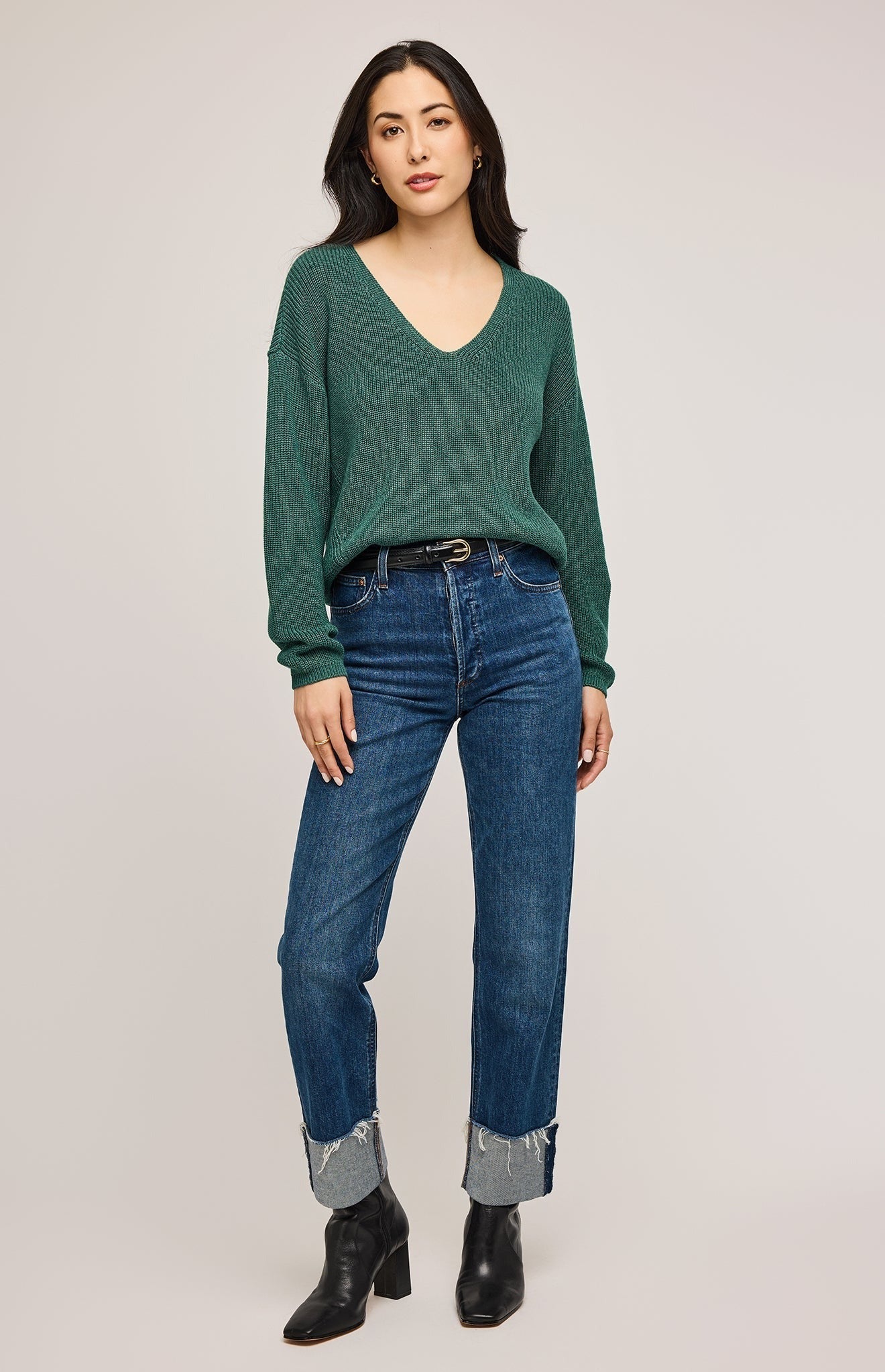 Femme qui porte un pull tricoté vert forêt avec un col en V associé avec un pantalon jeans indigo et un ceinture en cuir noir à boucle doré et des bottes à talons carrés en cuir noir. Vu en entier de face