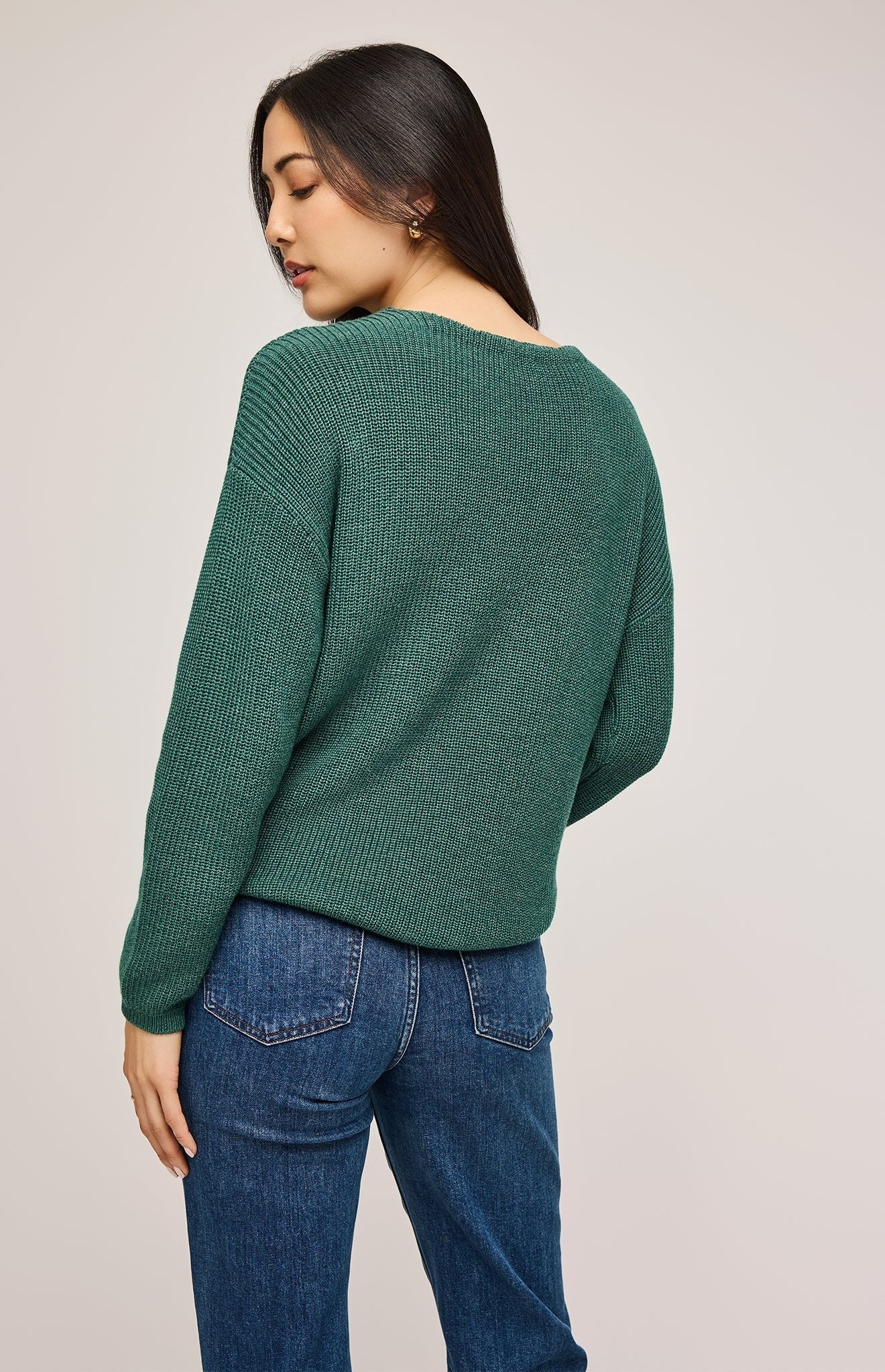 Femme qui porte un pull tricoté vert forêt avec un col en V associé avec un pantalon jeans indigo et un ceinture en cuir noir à boucle doré. Vu de dos