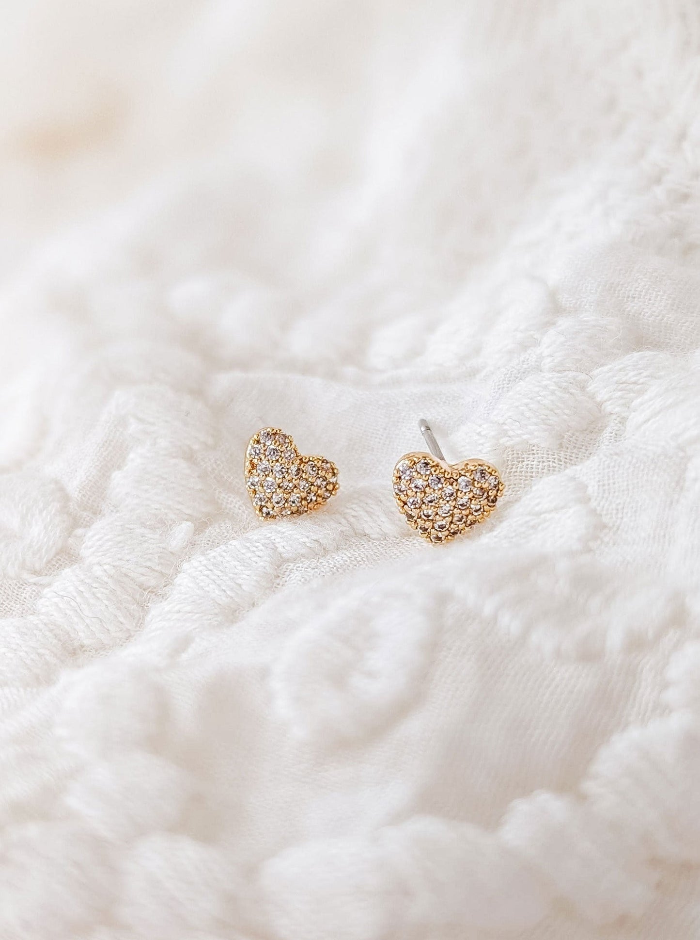 Petits clous d'oreilles dorés en forme de coeur ornées de zircons sur toute la surface. Les boucles d'oreilles sont déposées sur un tissu blanc. 