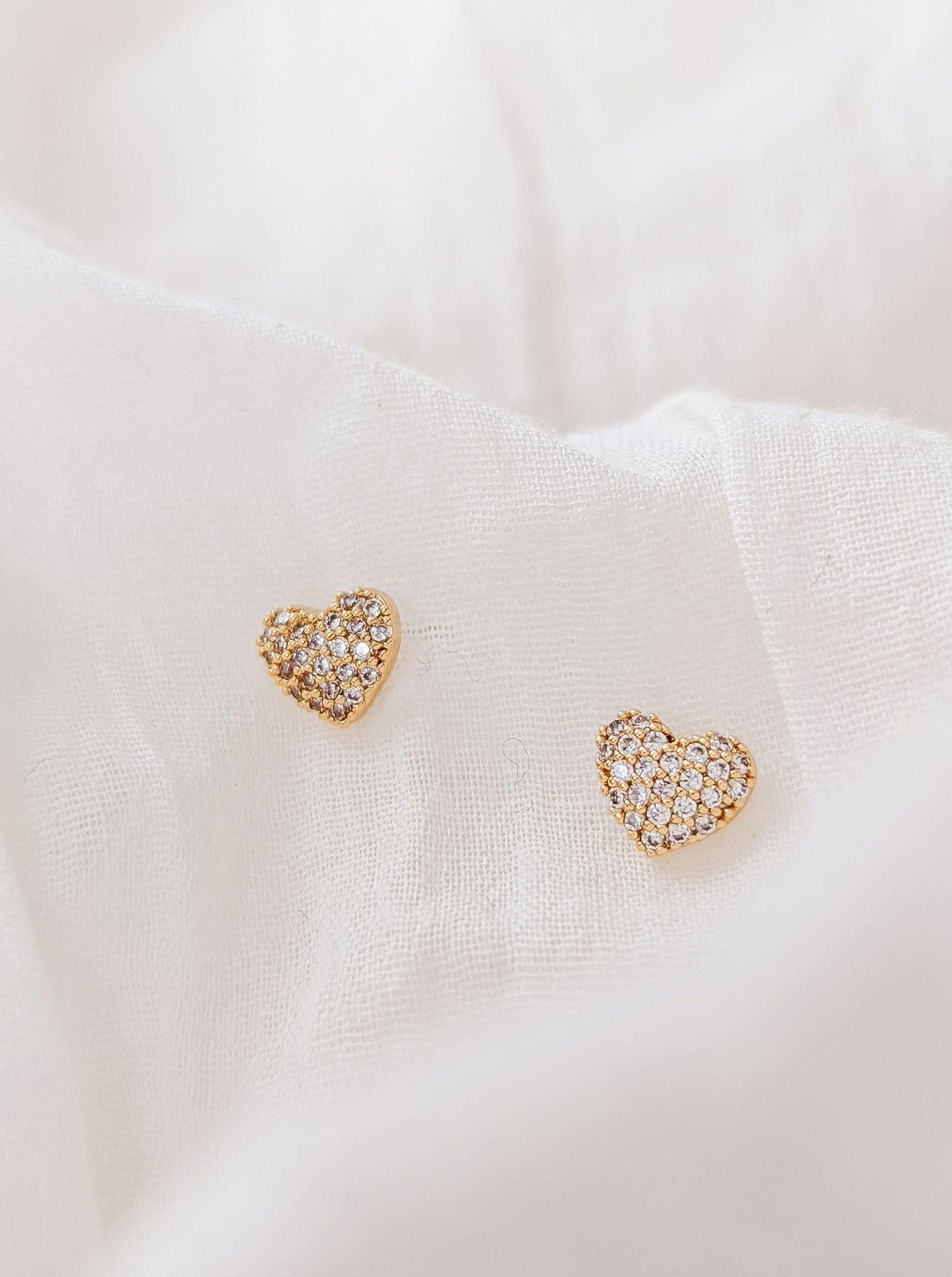 Petits clous d'oreilles dorés en forme de coeur ornées de zircons sur toute la surface. Les boucles d'oreilles sont déposées de face sur un tissu blanc. 