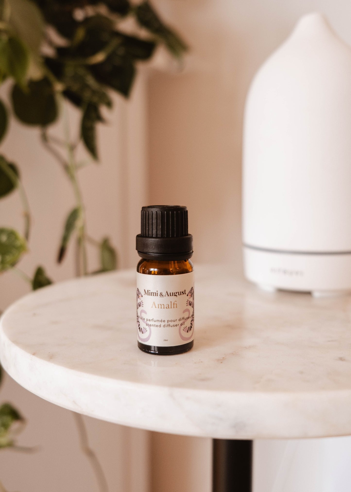 Le flacon d'huile parfumée est une petite bouteille en verre brun sur laquelle est collé une large étiquette beige. Dessus, on peut lire Mimi & August, Amalfi, huile parfumée pour diffuseur, scented diffuser oil. La bouteille est fermée, elle est déposée sur une table en marbre blanc, devant une plante et un diffuseur d'huile essentielle. 