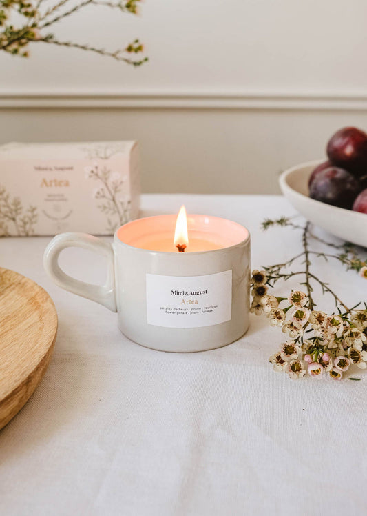 Tasse de taille moyenne en céramique beige avec une petite anse. Sur la tasse, il y a une étiquette blanche sur laquelle il est écrit Mimi & August, Artea et les notes de la bougie qui sont ici: pétales de rose, prune et feuillage. La bougie est déposé sur une table couverte d'une nappe beige, de fleurs séchée et de prunes. 