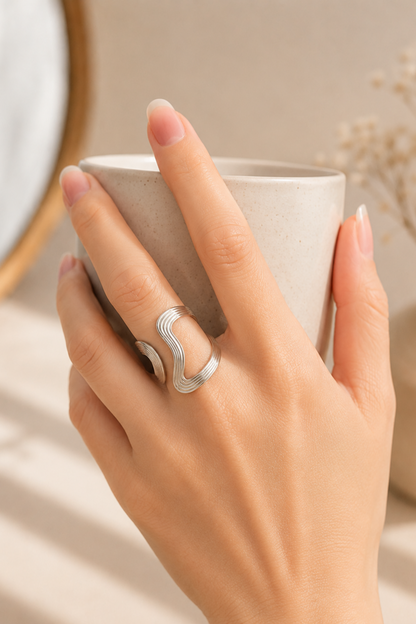 Bague Anna argent portée sur la main tenant une tasse, avec design ondulé ajustable et finition brillante.